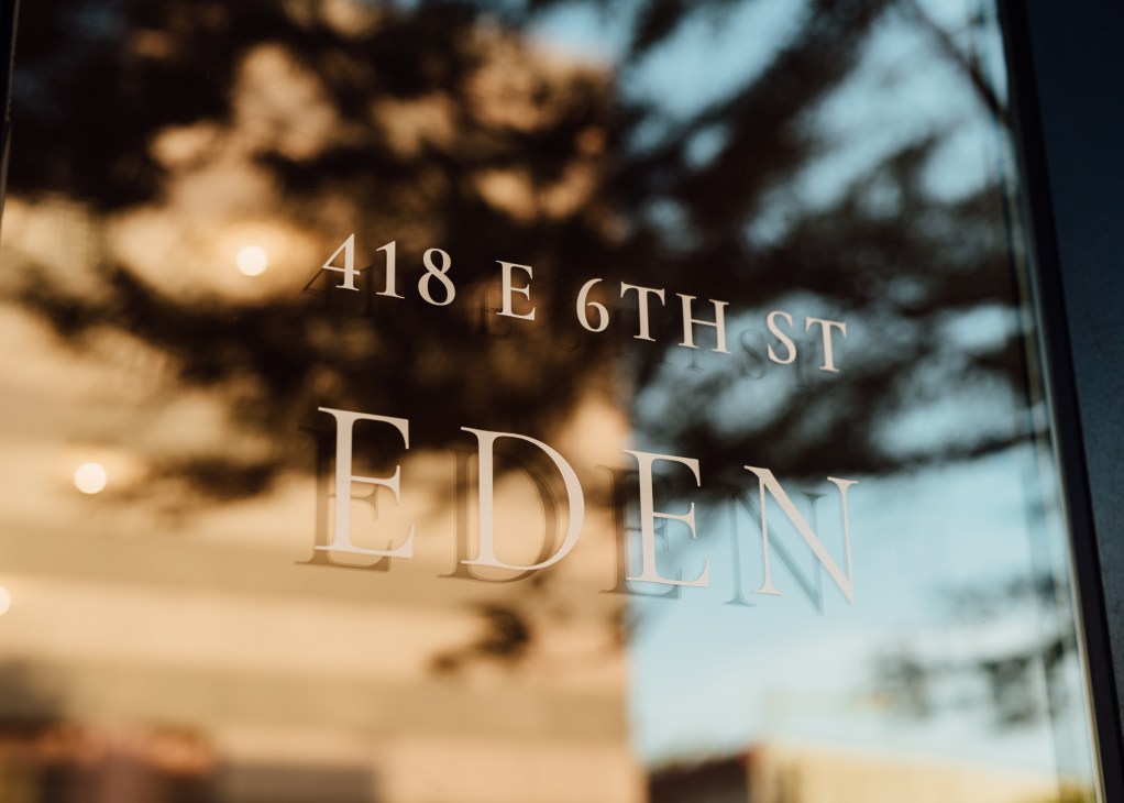 Glass storefront with etched address '418 E 6TH ST' and 'EDEN' text, showing tree reflections.
