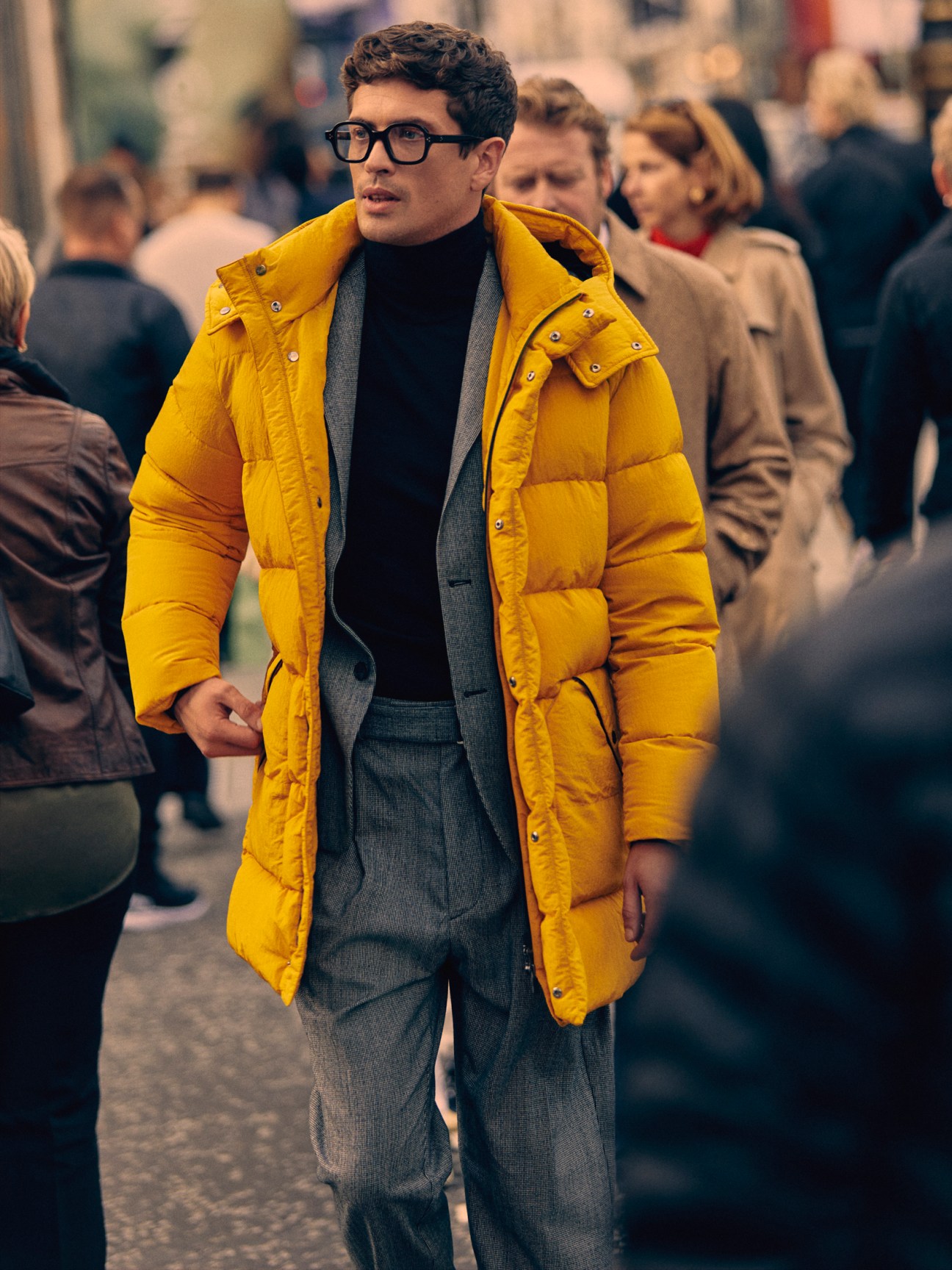 Man in bright yellow puffer coat over grey suit and black turtleneck walking through crowd