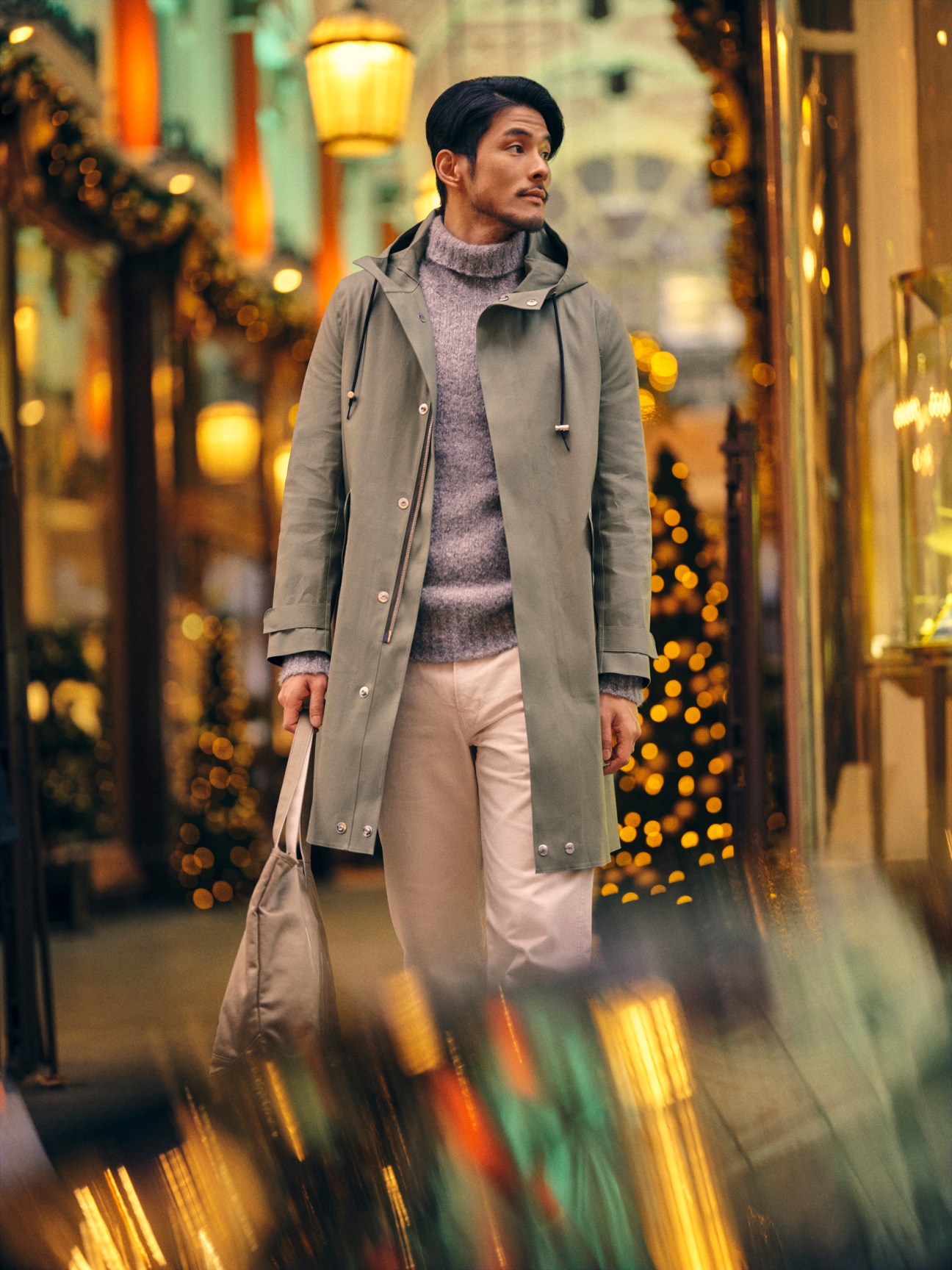 Man in beige coat and grey rollneck jumper carrying a bag on illuminated street