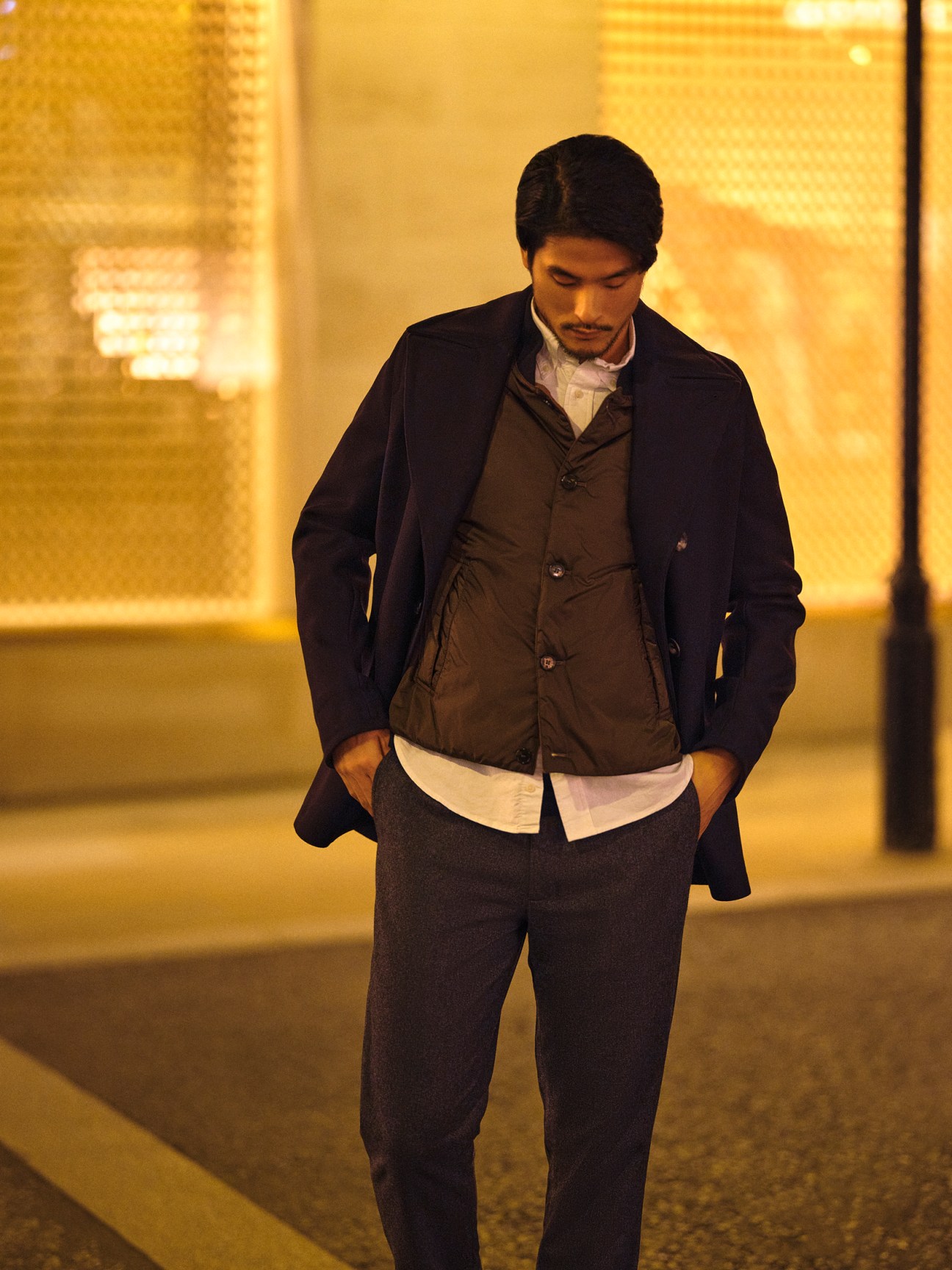 Man in light-colored coat and dark vest with white dress shirt walking outdoors at night