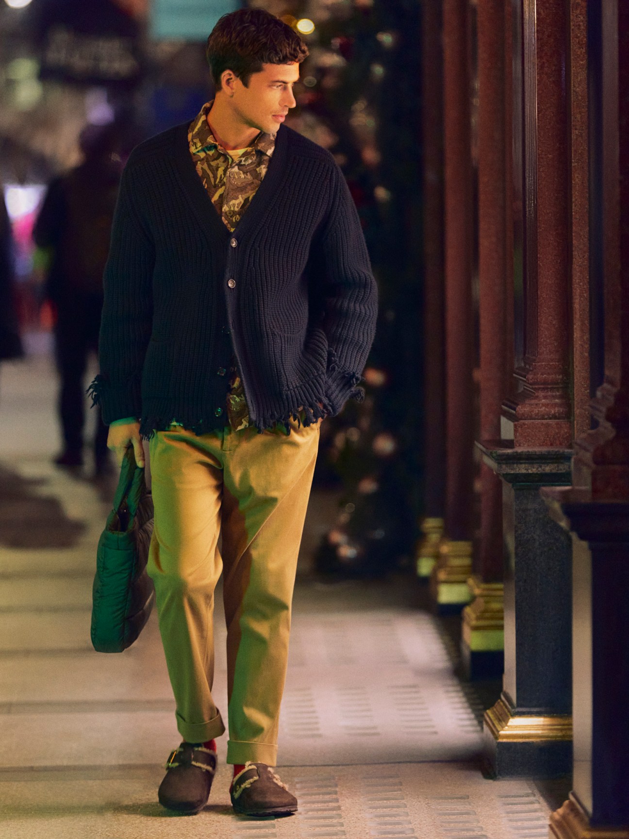 Man in cardigan and casual trousers wearing sandals with socks and carrying a bag