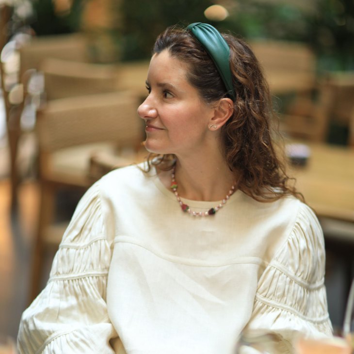 Woman with long brown hair wearing a cream-colored blouse and beaded necklace, seated indoors.