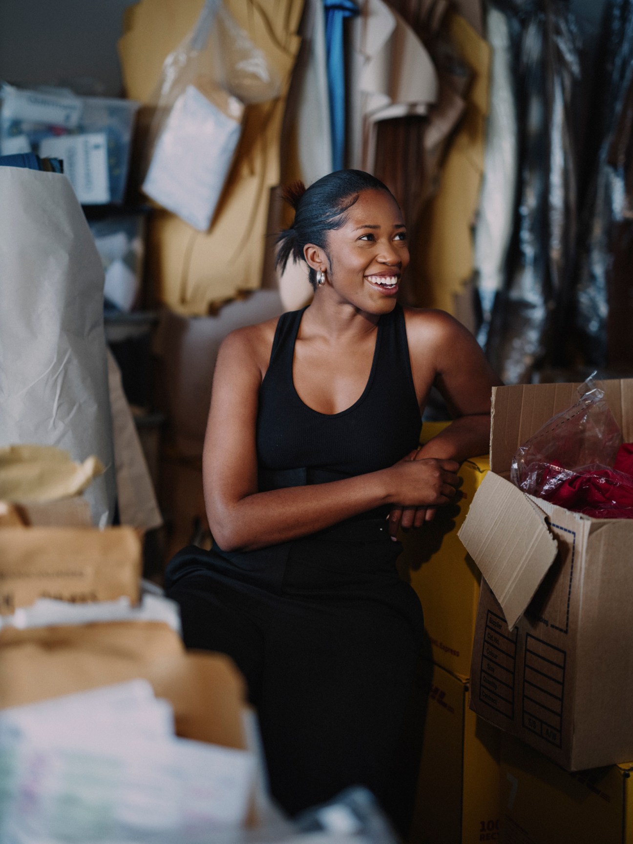 Bianca Saunders sitting among cardboard boxes and materials in her London studio workspace