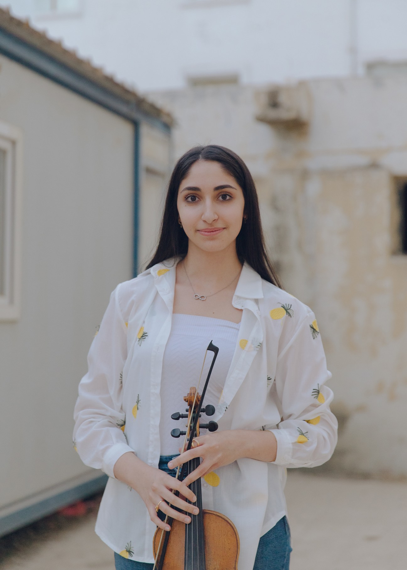 Young woman with long dark hair wearing white pineapple-print shirt holds violin outdoors