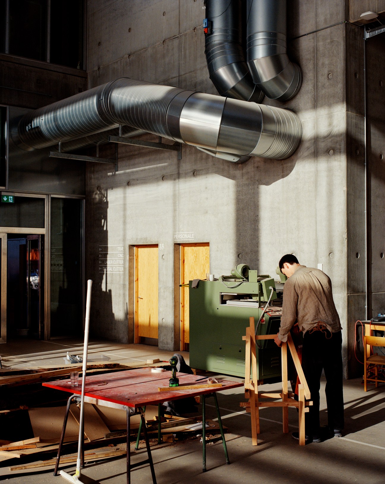 Student working at machinery in industrial wood workshop with ventilation ducts and concrete walls