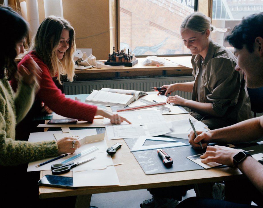 Students collaborating around a table with papers, books and materials in a bright studio space