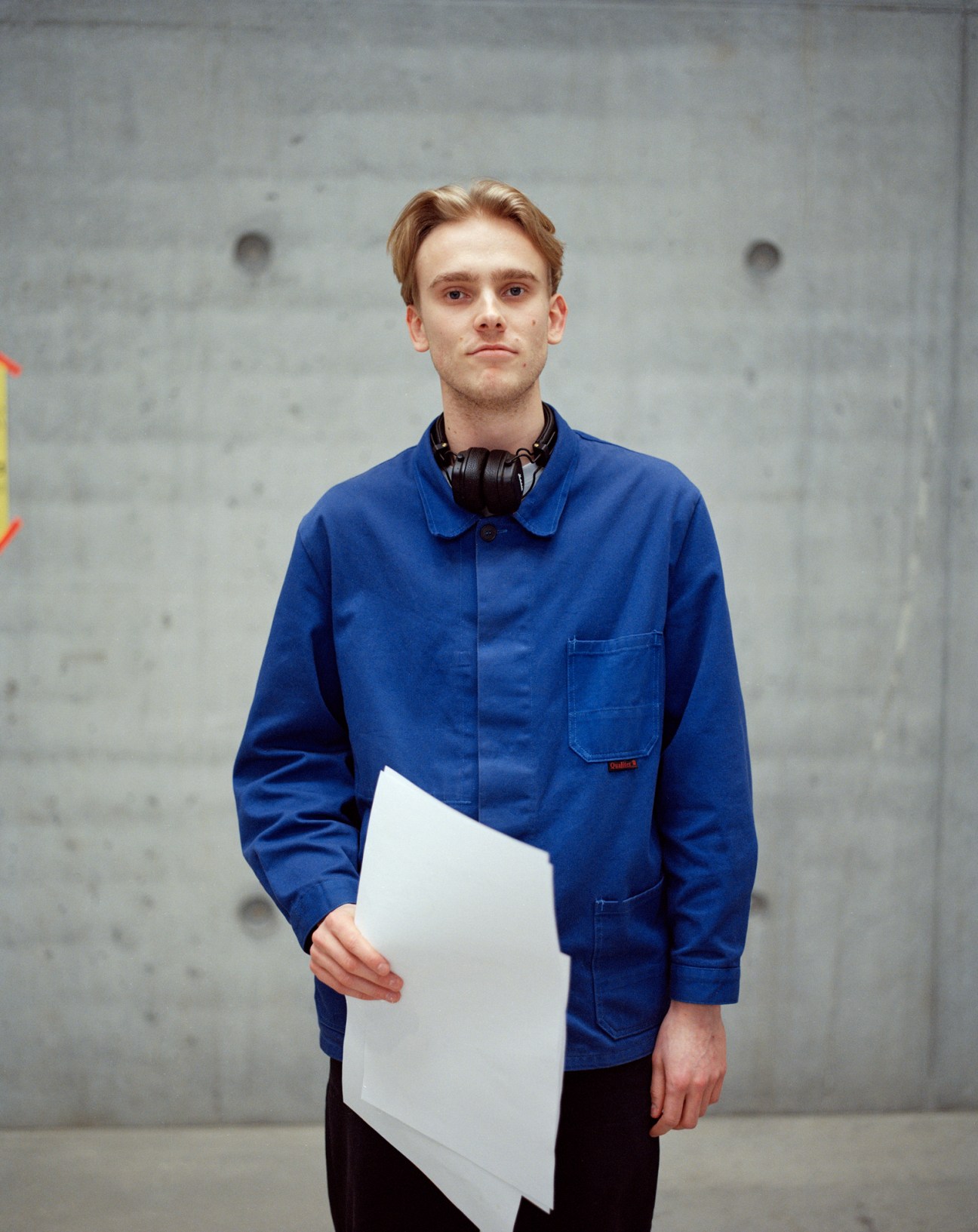 Bertram Øe, BA architecture student, wearing blue work jacket and headphones, holding white papers in concrete building