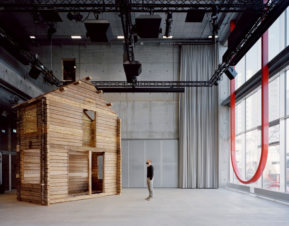 Wooden log cabin structure displayed in large industrial space with concrete walls and exposed ceiling