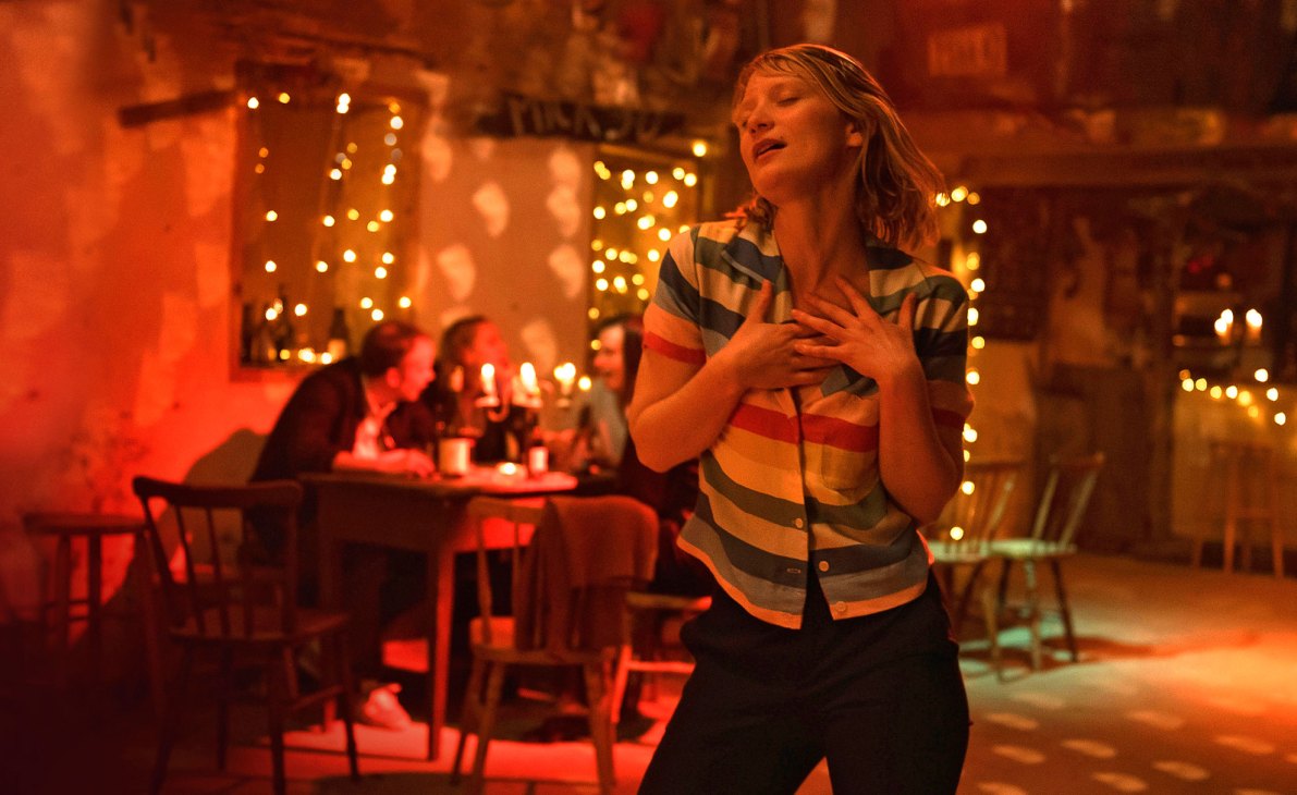 Woman with hand on chest in warmly lit restaurant or bar with string lights and blurred diners in background