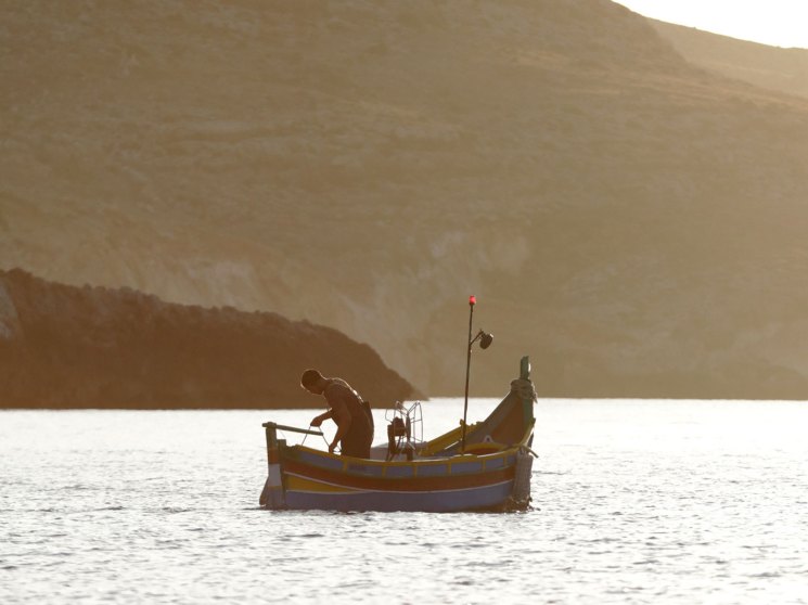Fisherman working on small yellow boat on calm water with mountainous coastline in golden light