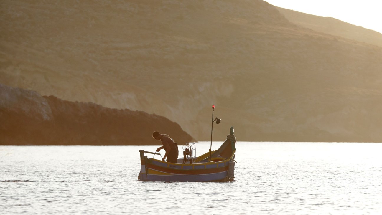 Fisherman working on small yellow boat on calm water with mountainous coastline in golden light