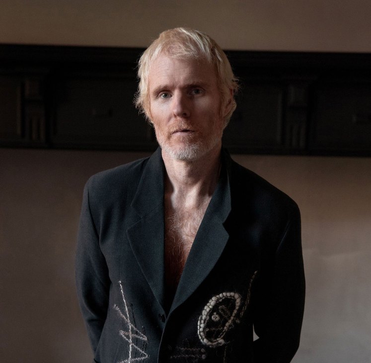 Man with blonde hair and beard wearing dark blazer with decorative embellishments in interior setting