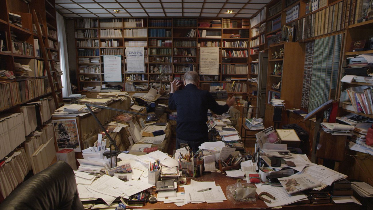 Elderly man sits amid chaotic study filled with scattered papers, books, and musical scores covering every surface