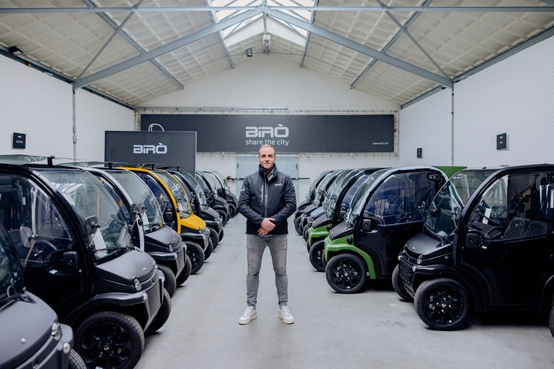 Man in dark jacket standing between rows of small electric vehicles in warehouse with Birò branding