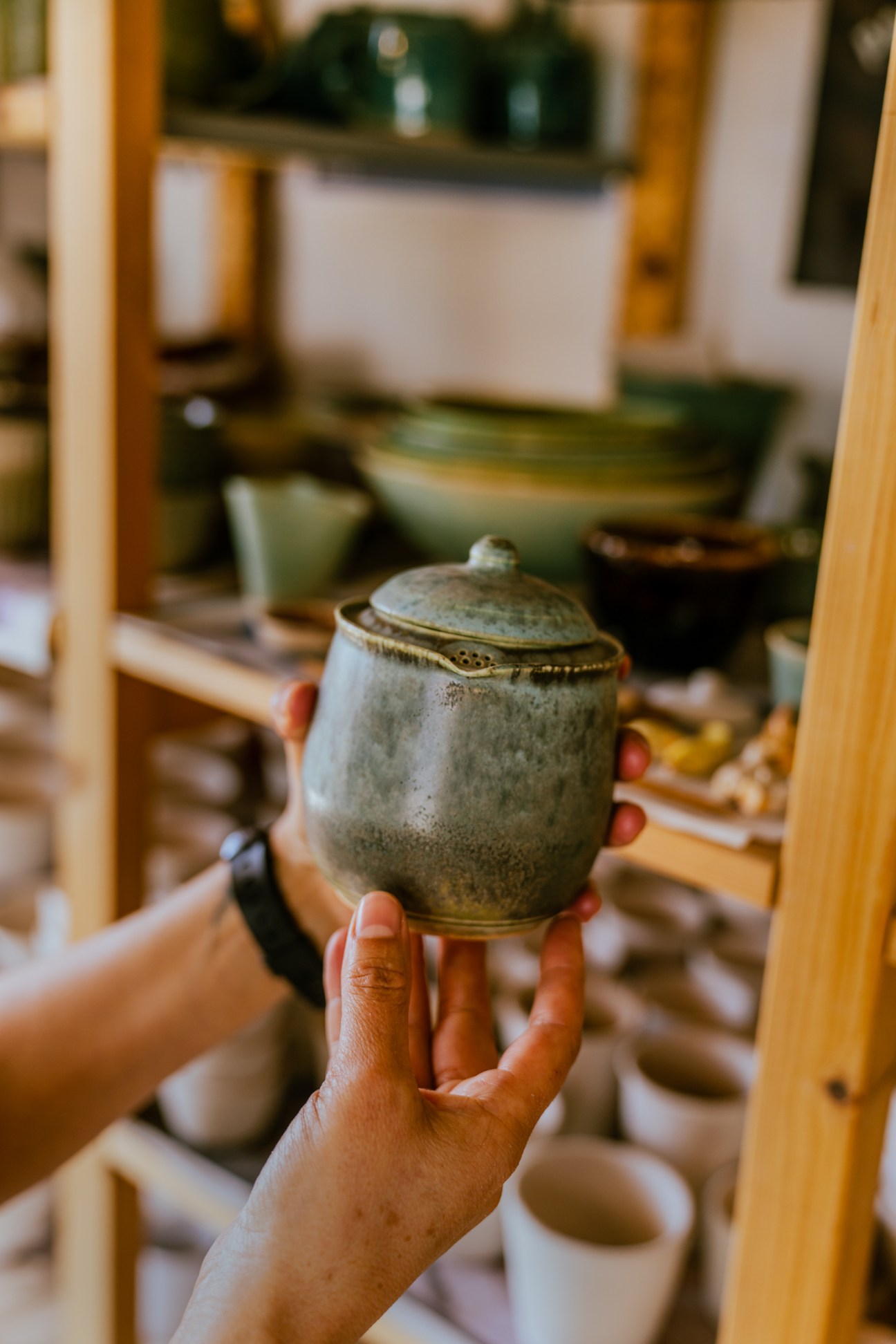 Hands holding a handmade ceramic jar with lid by Adriana Berumen in pottery workshop with shelves of ceramics