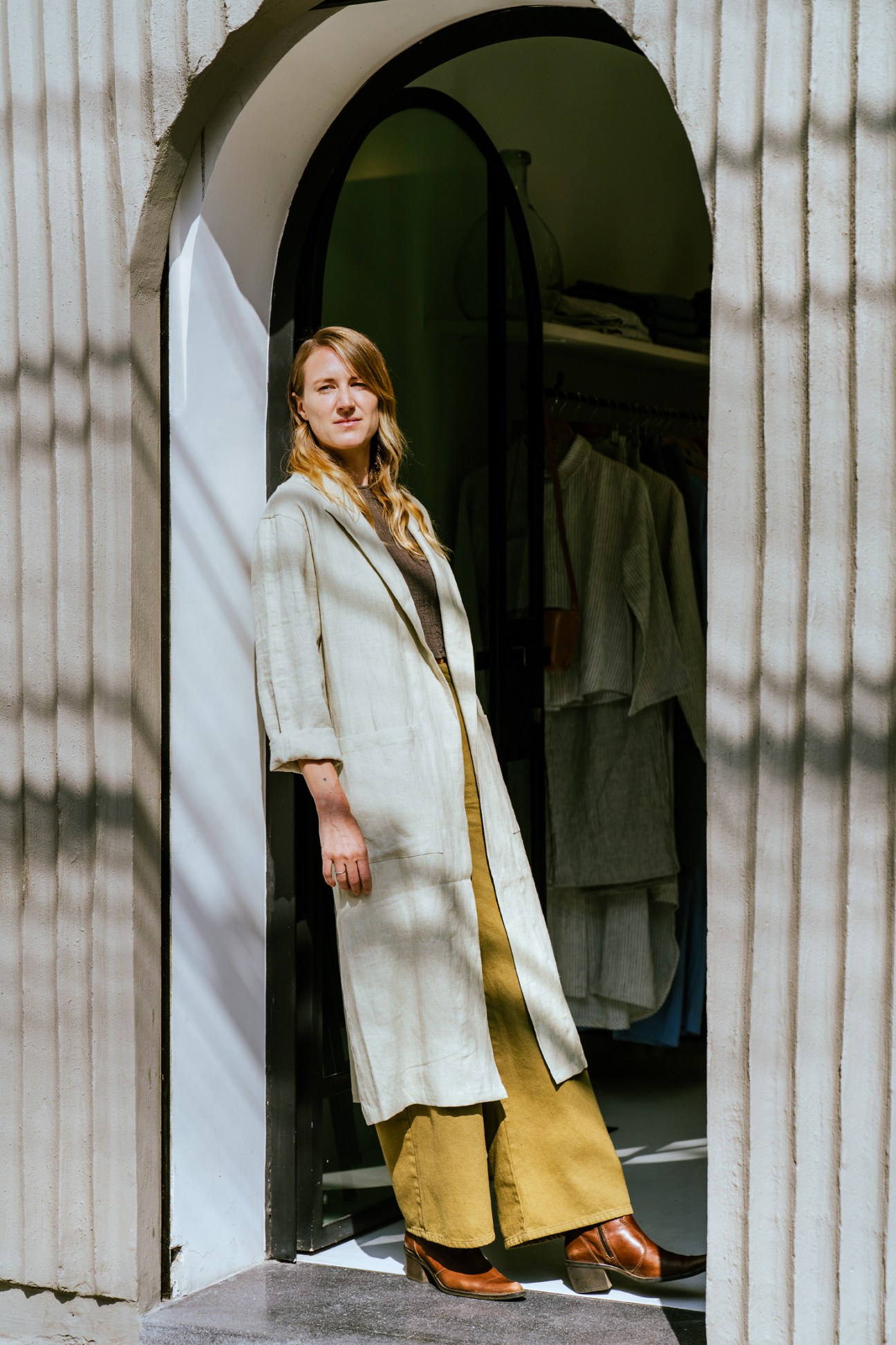 Bridget Tidey standing in doorway wearing cream coat and yellow pants at her clothing store Zii Ropa