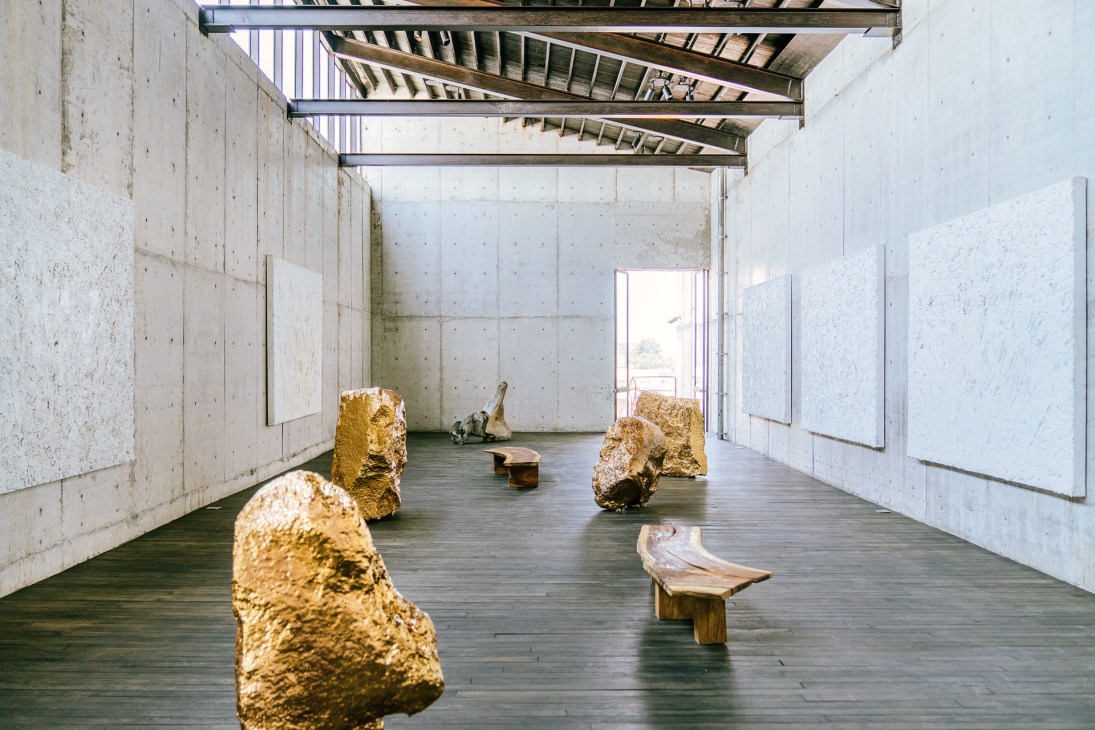Interior gallery space with gold-coated spheres and wooden sculpture displayed in minimalist concrete room with skylights.