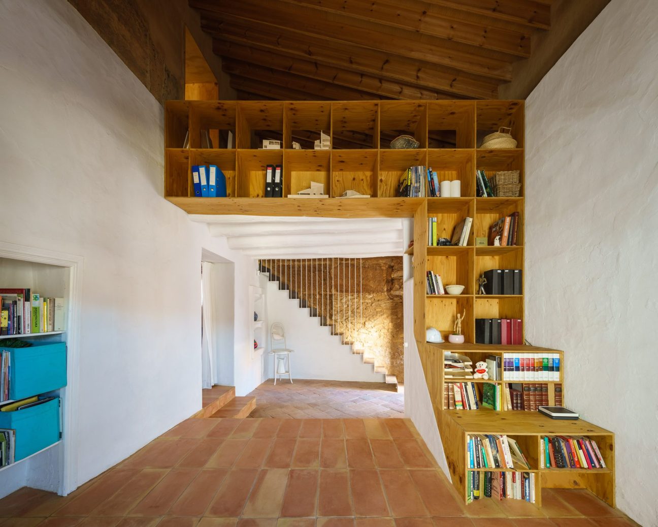 Modern interior with wooden ceiling beams, built-in shelving unit filled with books, terracotta tile flooring and white staircase