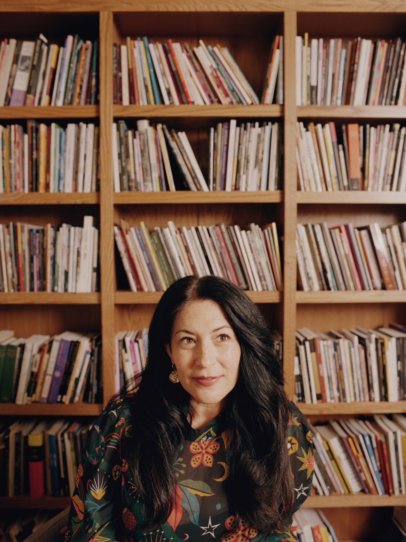 Ada Limón, the US poet laureate a woman with long dark hair wearing floral patterned shirt sits in front of wooden bookshelf filled with books