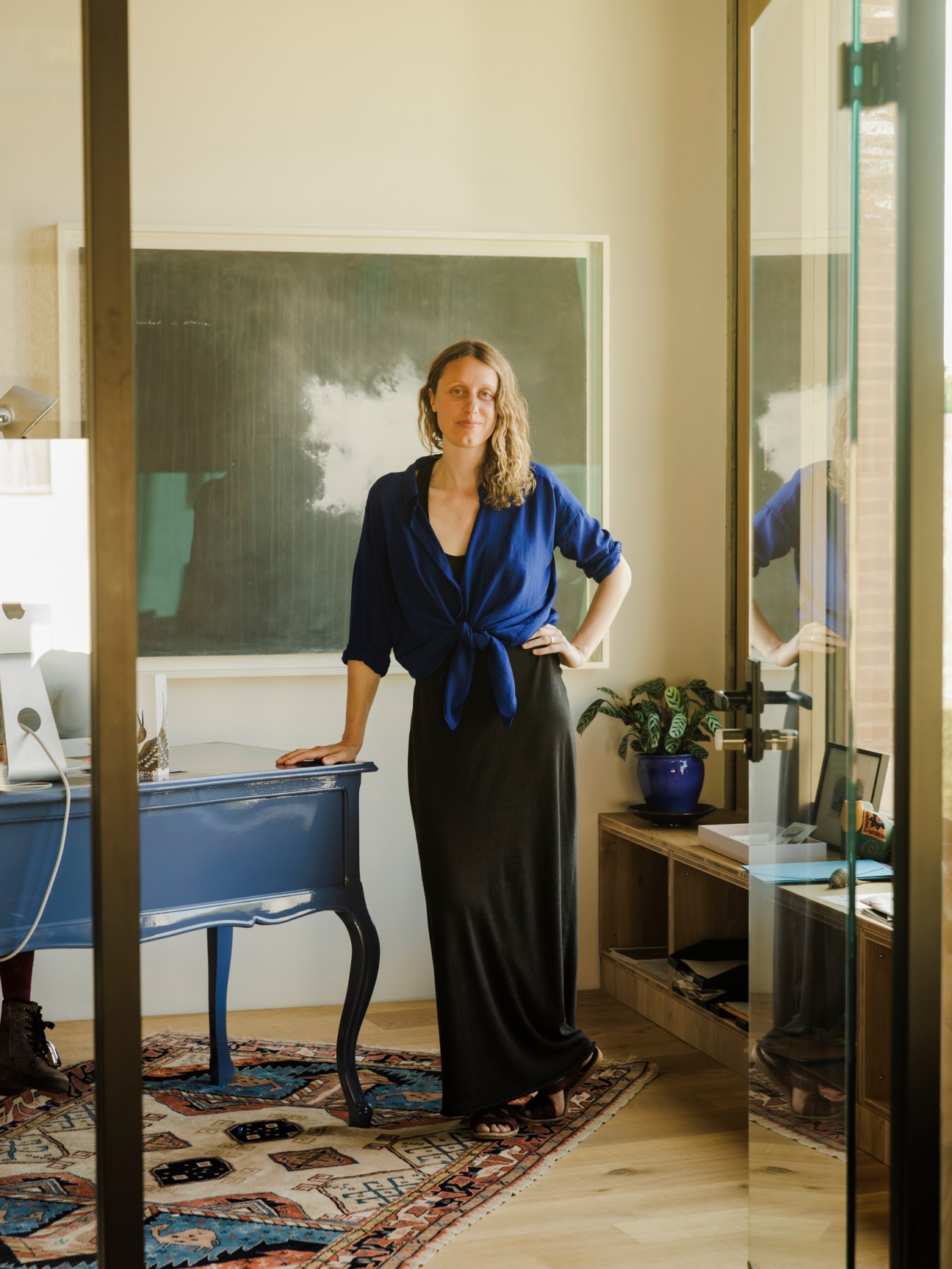 Woman in blue shirt standing in modern interior with artwork, plants, and wooden furniture