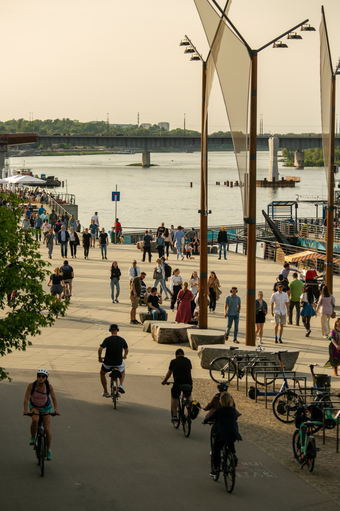 People walking and cycling along Warsaw's Vistula riverbank with modern architectural structures and bridges visible