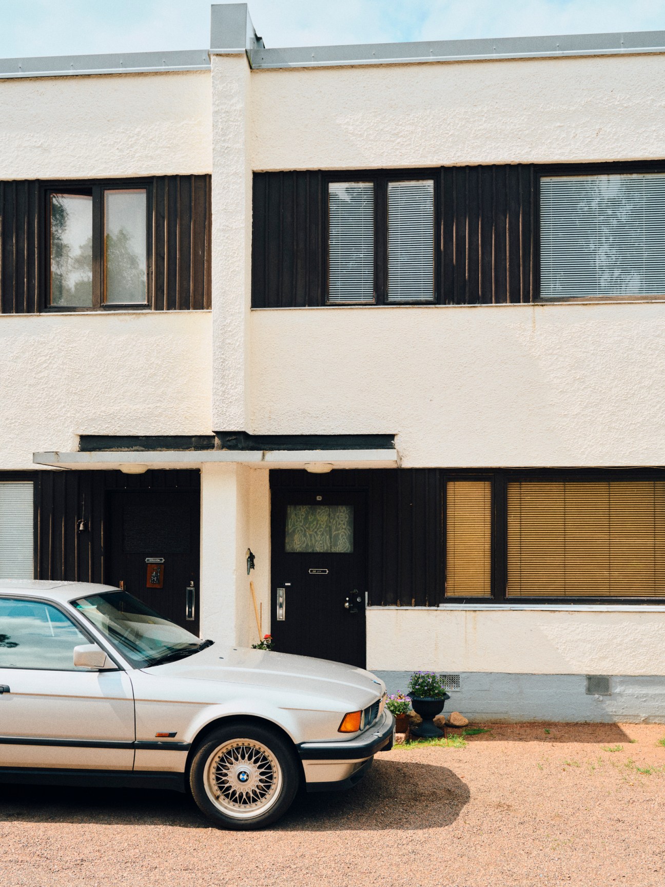 Cream-colored modernist terraced housing with dark wood shutters and a vintage BMW parked outside