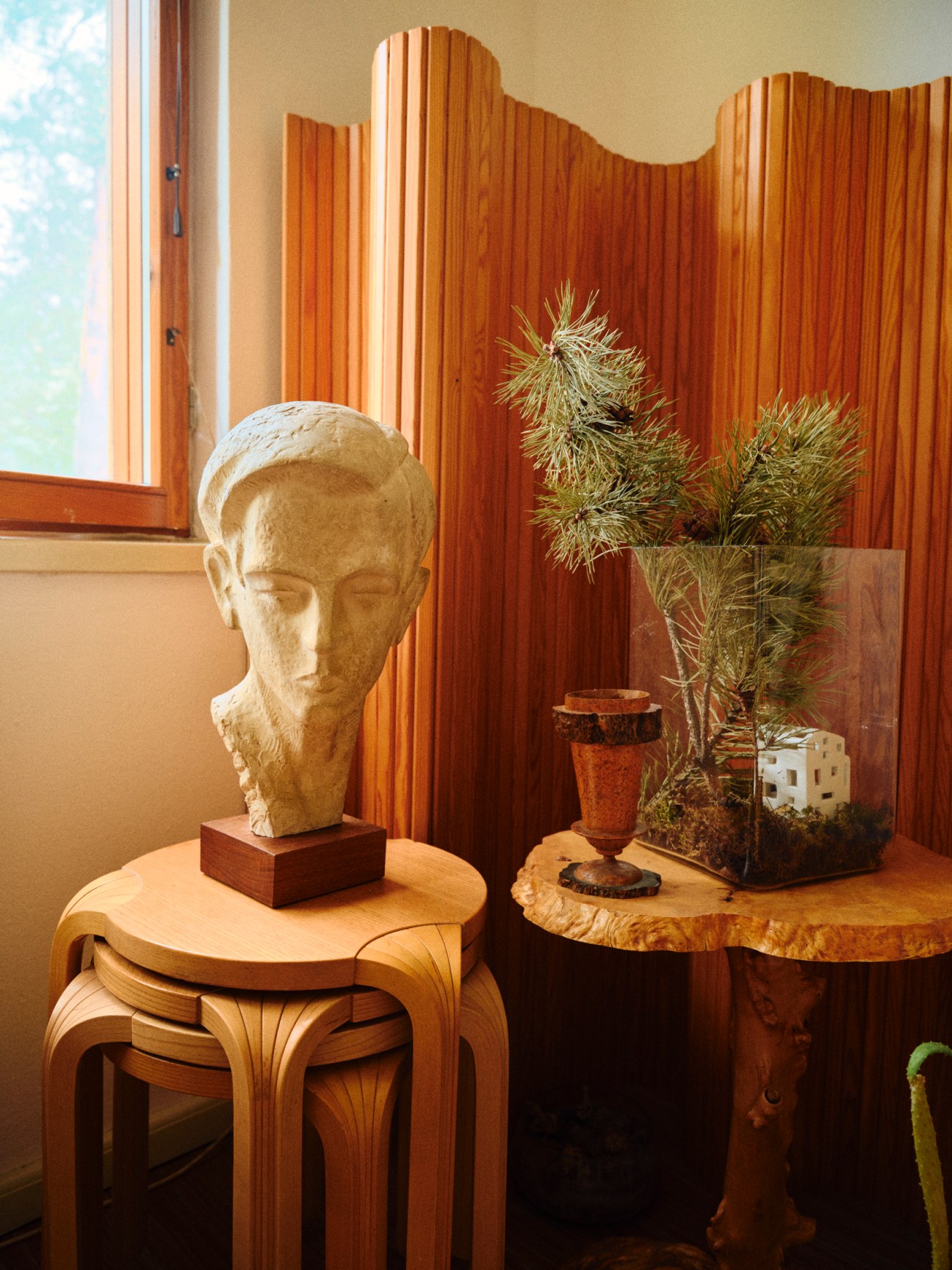 Classical sculpture bust on wooden stool next to plant and decorative objects in warm wooden interior