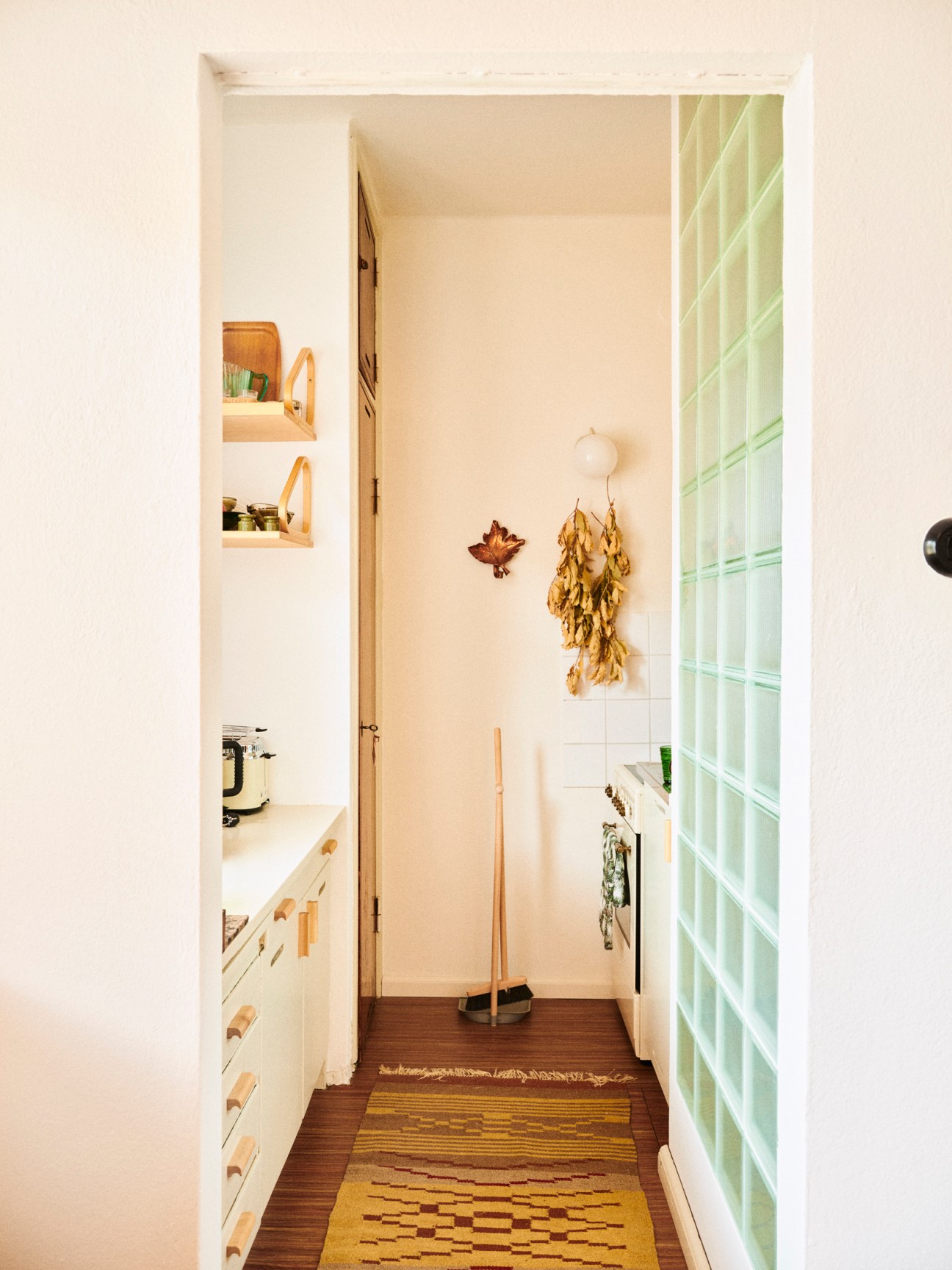 Narrow white kitchen pantry with built-in shelving, dried herbs hanging on wall, and green glass block window