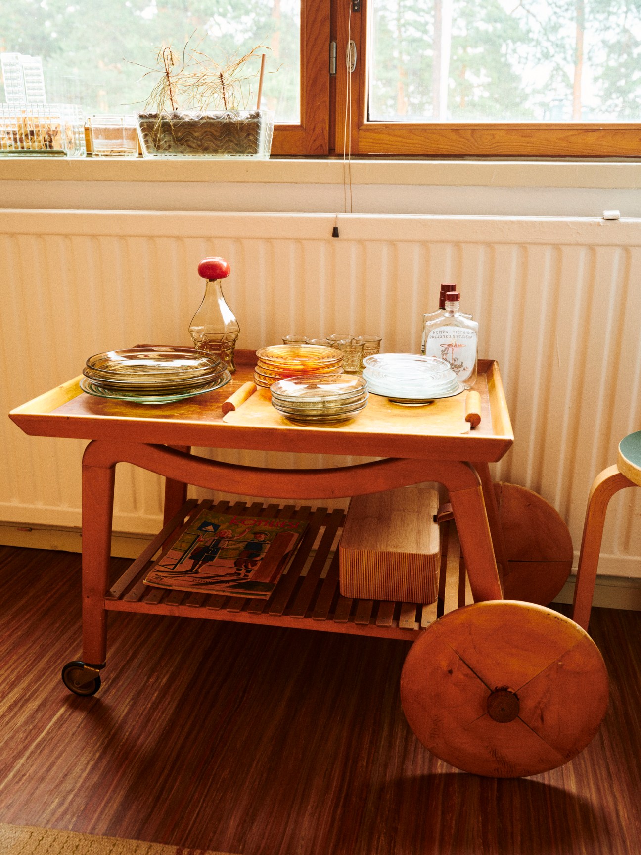 A vintage wooden bar trolley with round wheels holds stacked plates, glassware and bottles in a sunlit room
