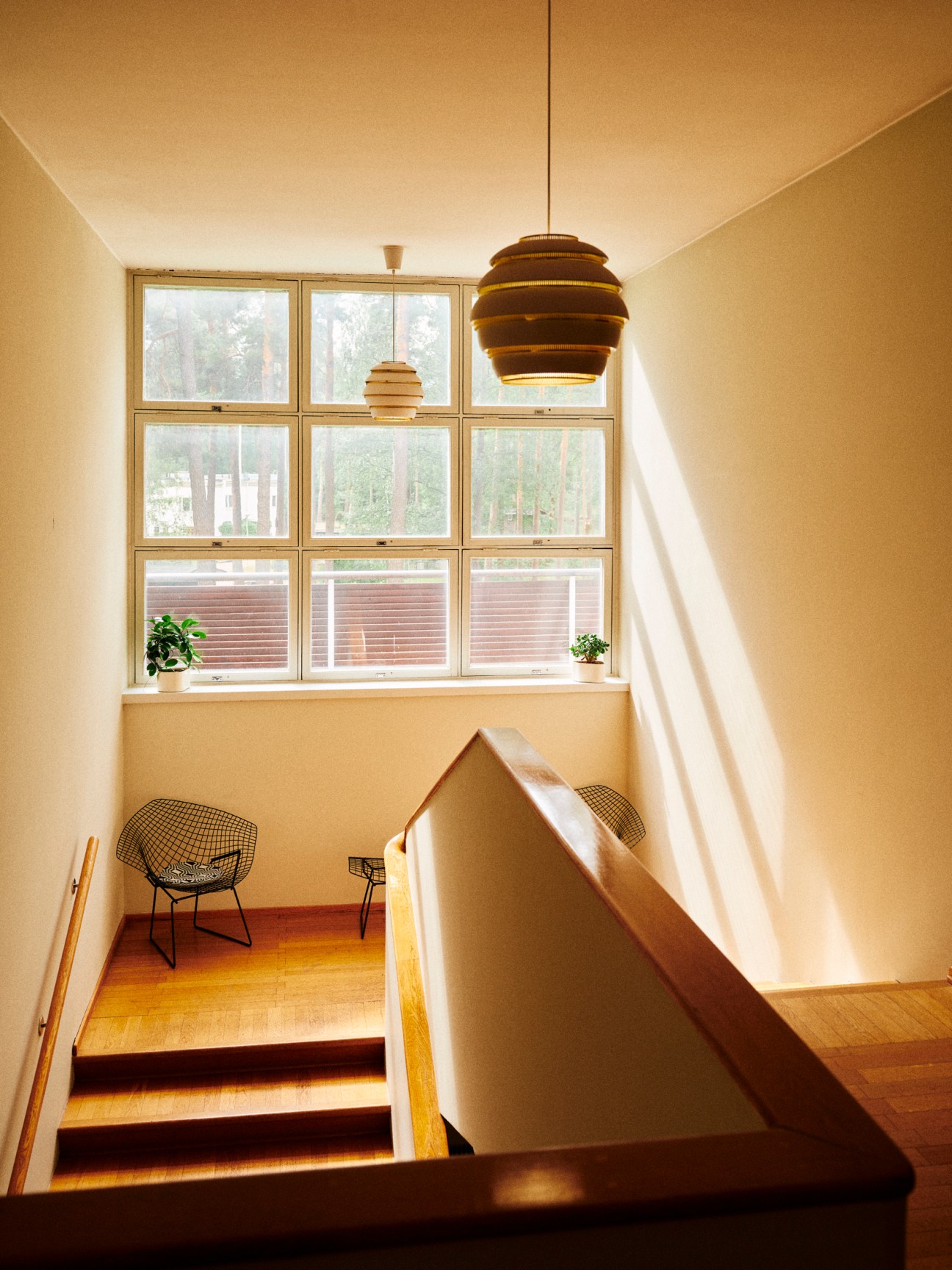 Bright staircase landing with modernist pendant light, wire chairs, and large grid windows overlooking forest