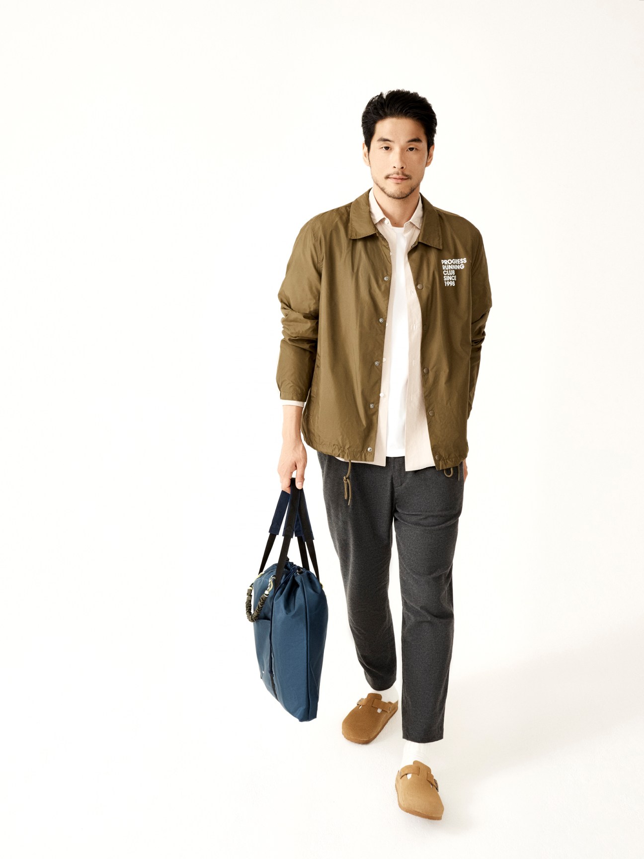 Asian man in olive jacket and dark trousers holding blue bag, wearing brown sandals on white background