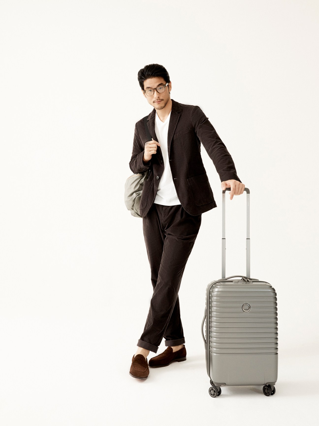 Man in dark suit with glasses holding backpack and standing next to silver rolling suitcase on white background