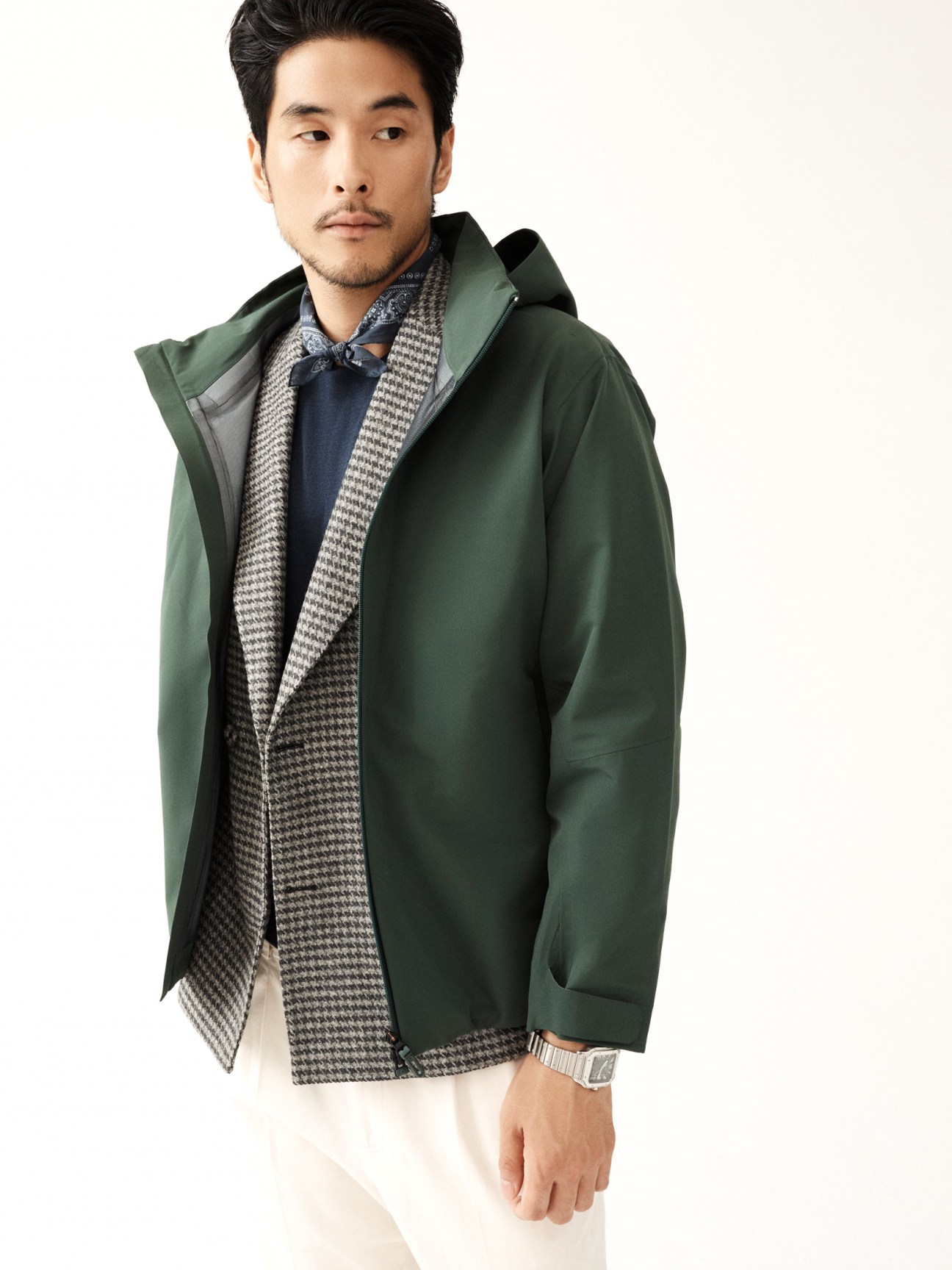 Asian man in green hooded coat layered over houndstooth jacket and navy sweater against white background