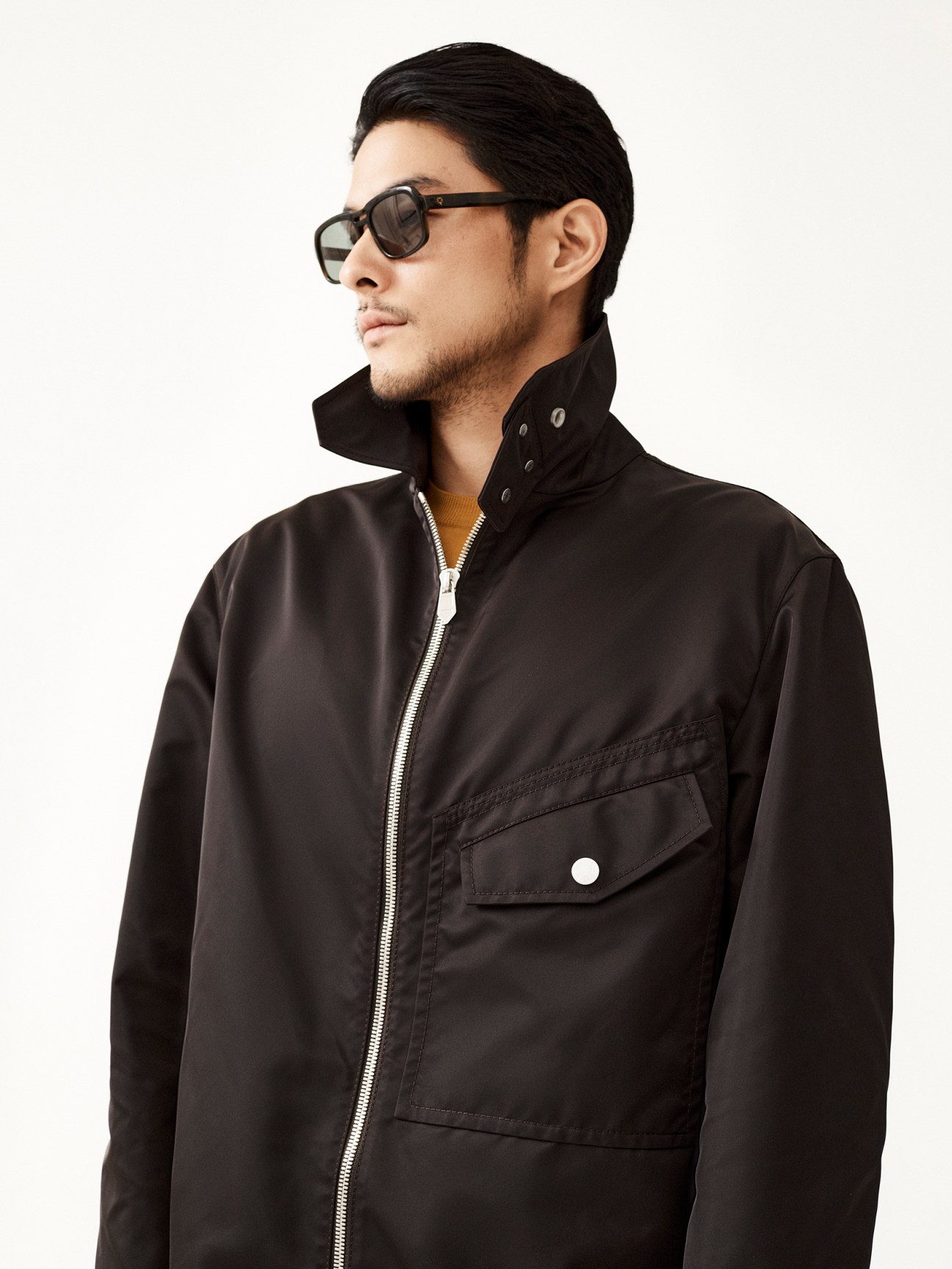 Man in profile wearing black zip-up jacket with chest pocket, orange sweater underneath, and dark sunglasses