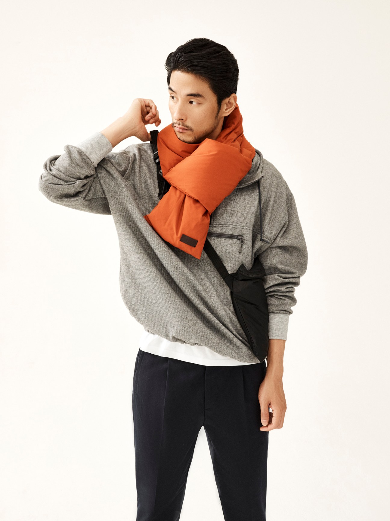 Asian man in gray sweatshirt and orange scarf poses with hand to chin against white background