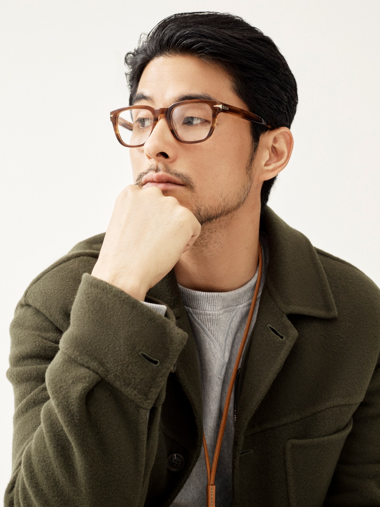 Asian man in olive green wool coat and brown-framed glasses poses thoughtfully with hand to chin