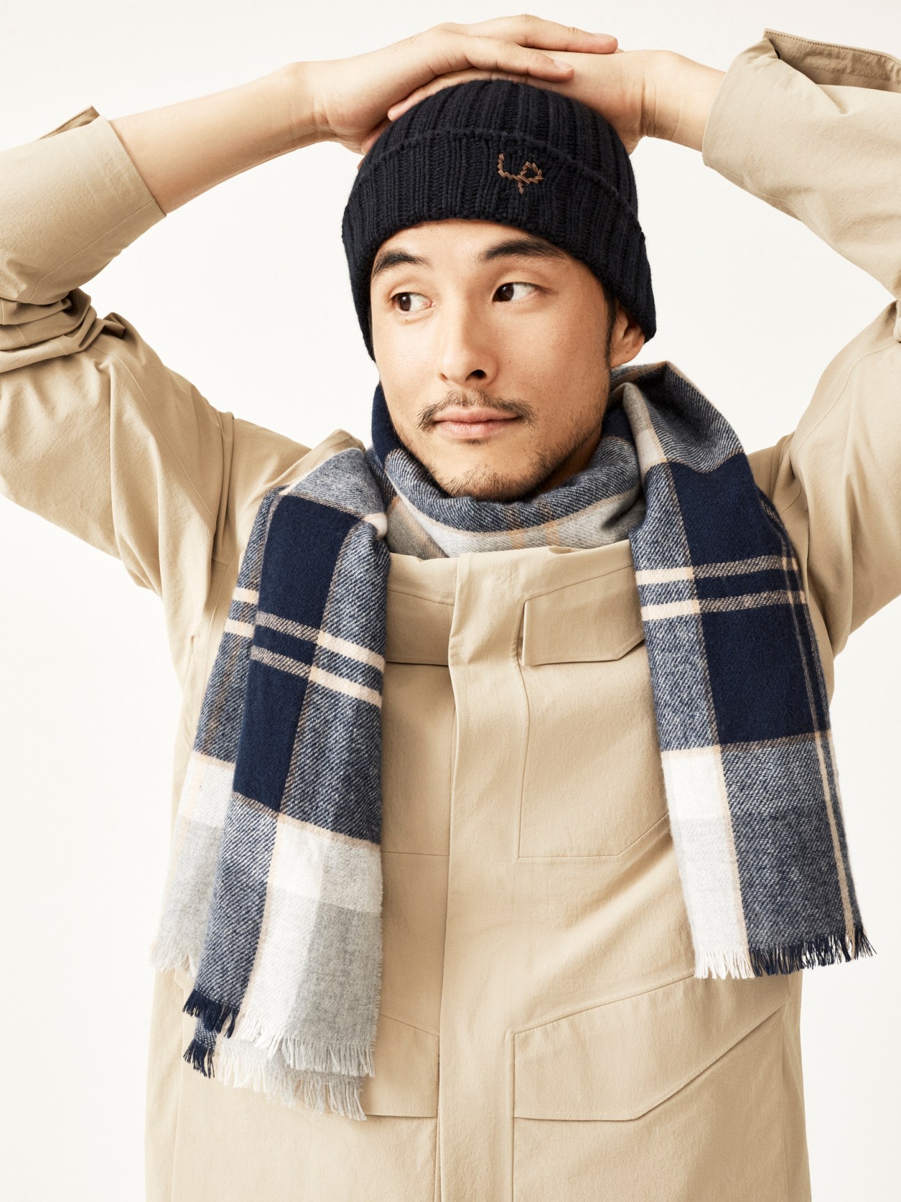 Asian man in beige jacket, navy beanie, and plaid scarf with hands positioned behind his head against white background