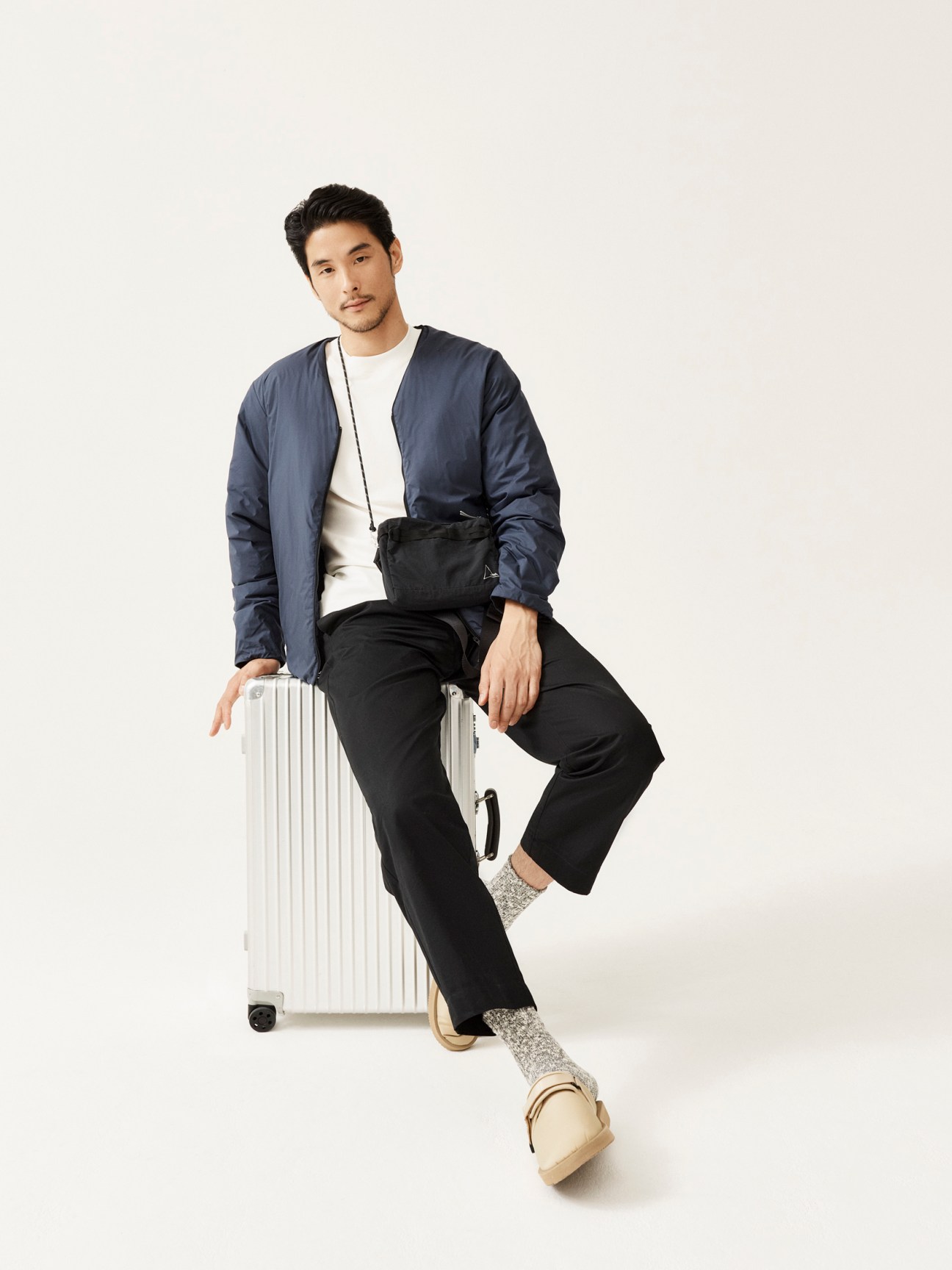 Man in navy jacket and black pants sitting on white suitcase with crossbody bag and sandals