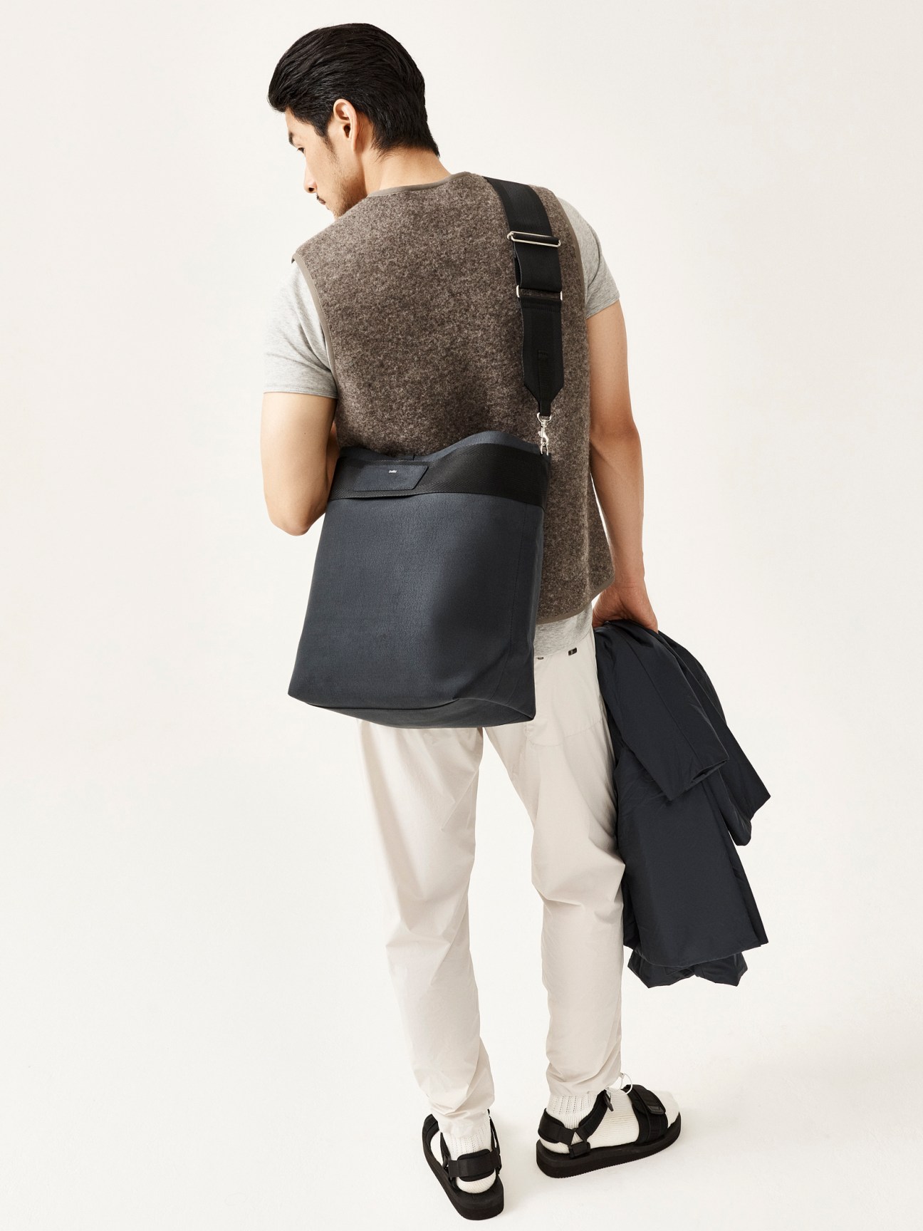 Man in layered outfit with grey vest, white pants and black sandals, carrying a dark jacket and crossbody bag