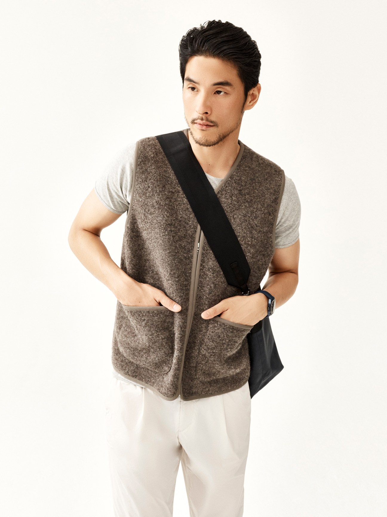 Asian man in gray knit vest over light t-shirt and white pants with black crossbody bag against white background