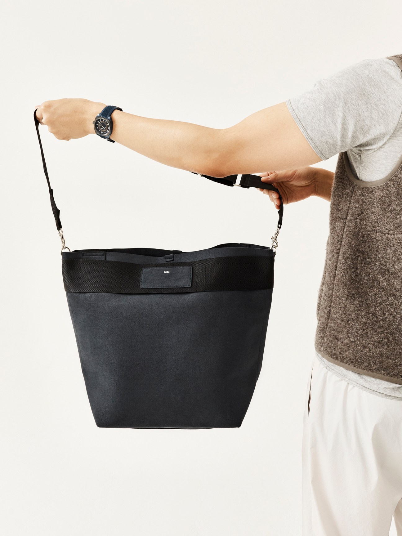 Person wearing grey sweater and white pants holding a black shoulder bag with adjustable strap