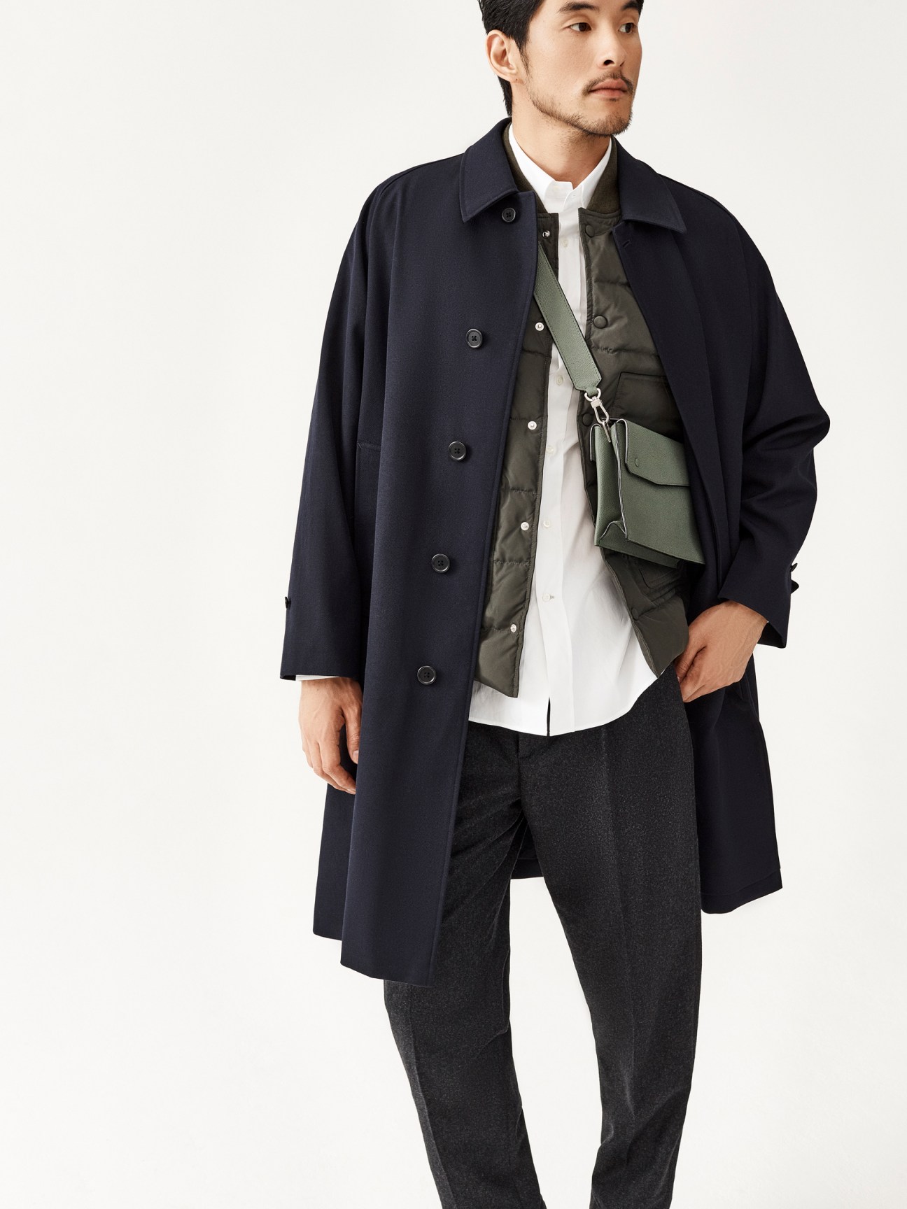 Asian man in navy coat over olive vest and white shirt with green crossbody bag and dark trousers
