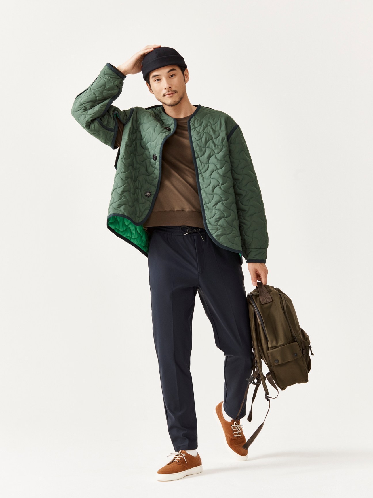 Man in green quilted jacket, brown shirt, navy trousers and beanie poses with olive backpack against white background
