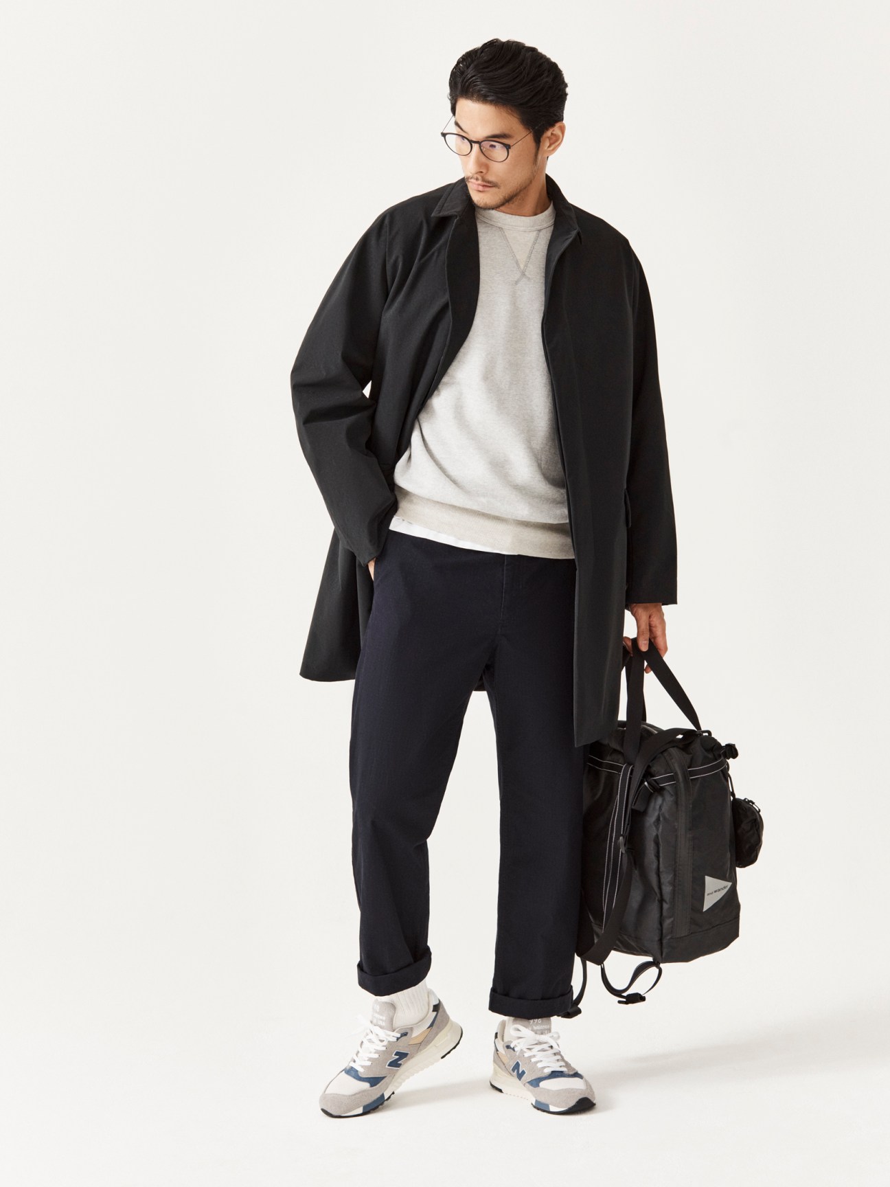 Man in black coat, grey sweatshirt, and dark trousers holding a black bag against white background