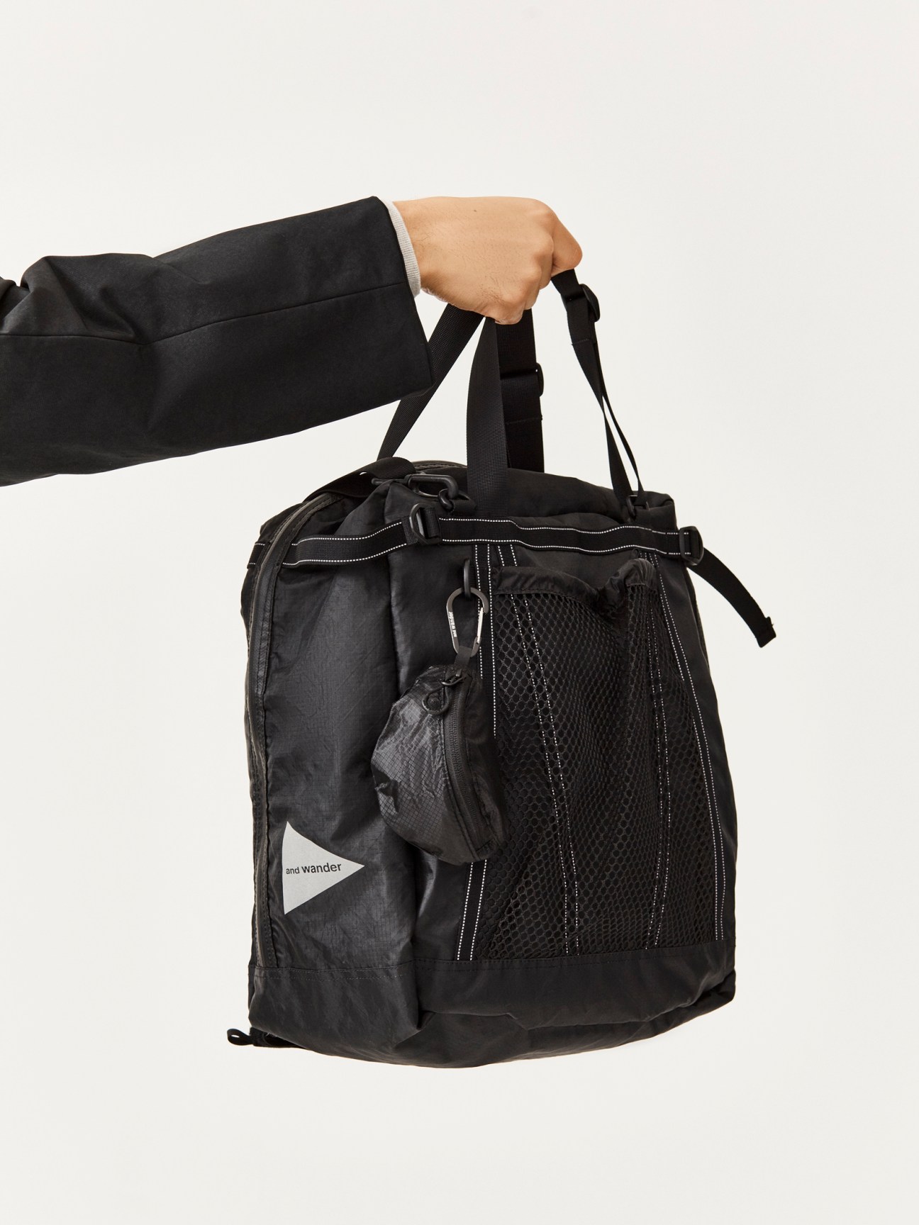 Person in black suit jacket holding a black And Wander backpack with mesh side pockets and white logo patch