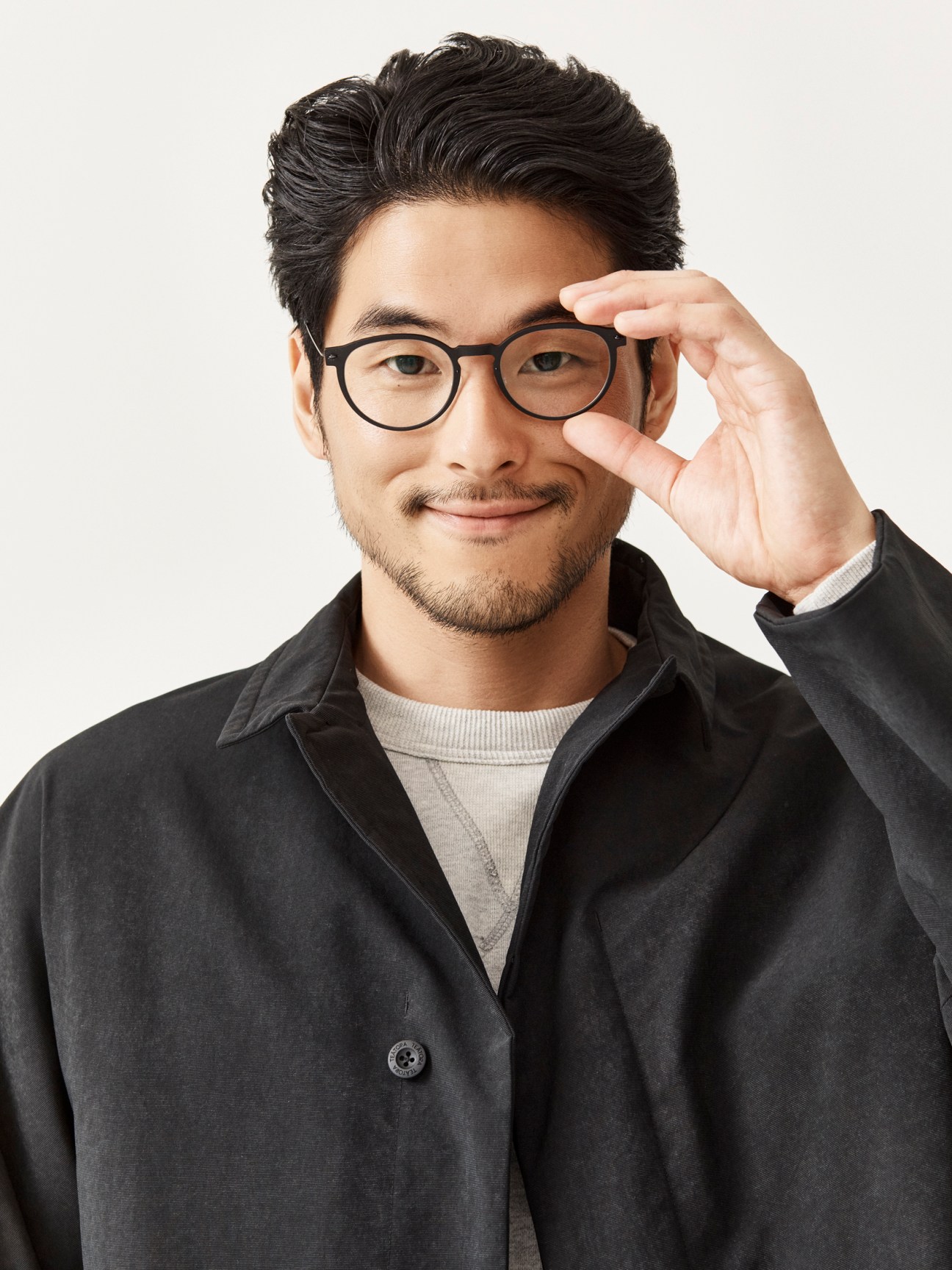 Asian man with glasses and facial hair wearing dark button-up shirt over beige sweater, hand raised to glasses