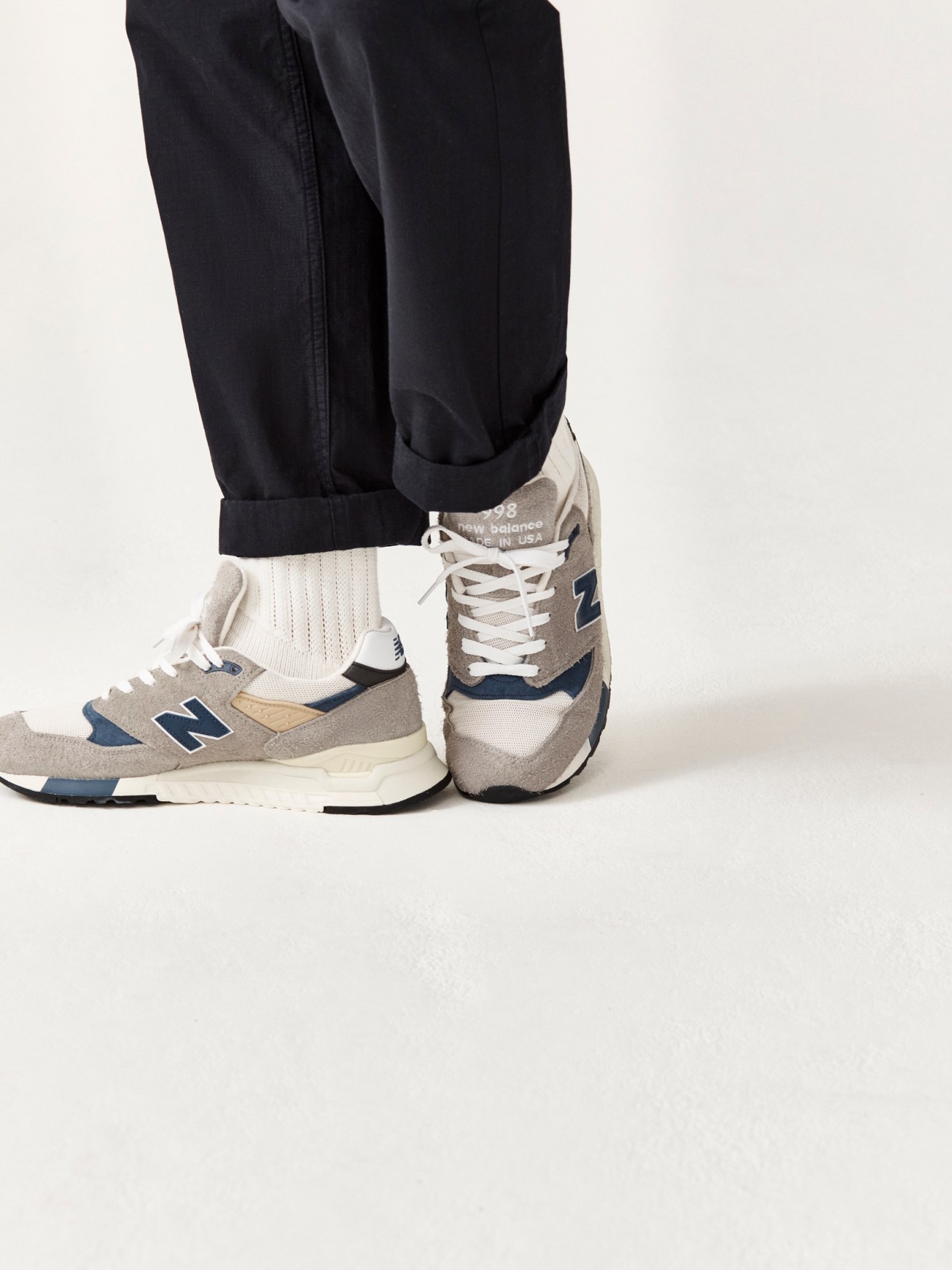 Person wearing black rolled-up trousers, white ribbed socks, and grey New Balance sneakers with blue accents