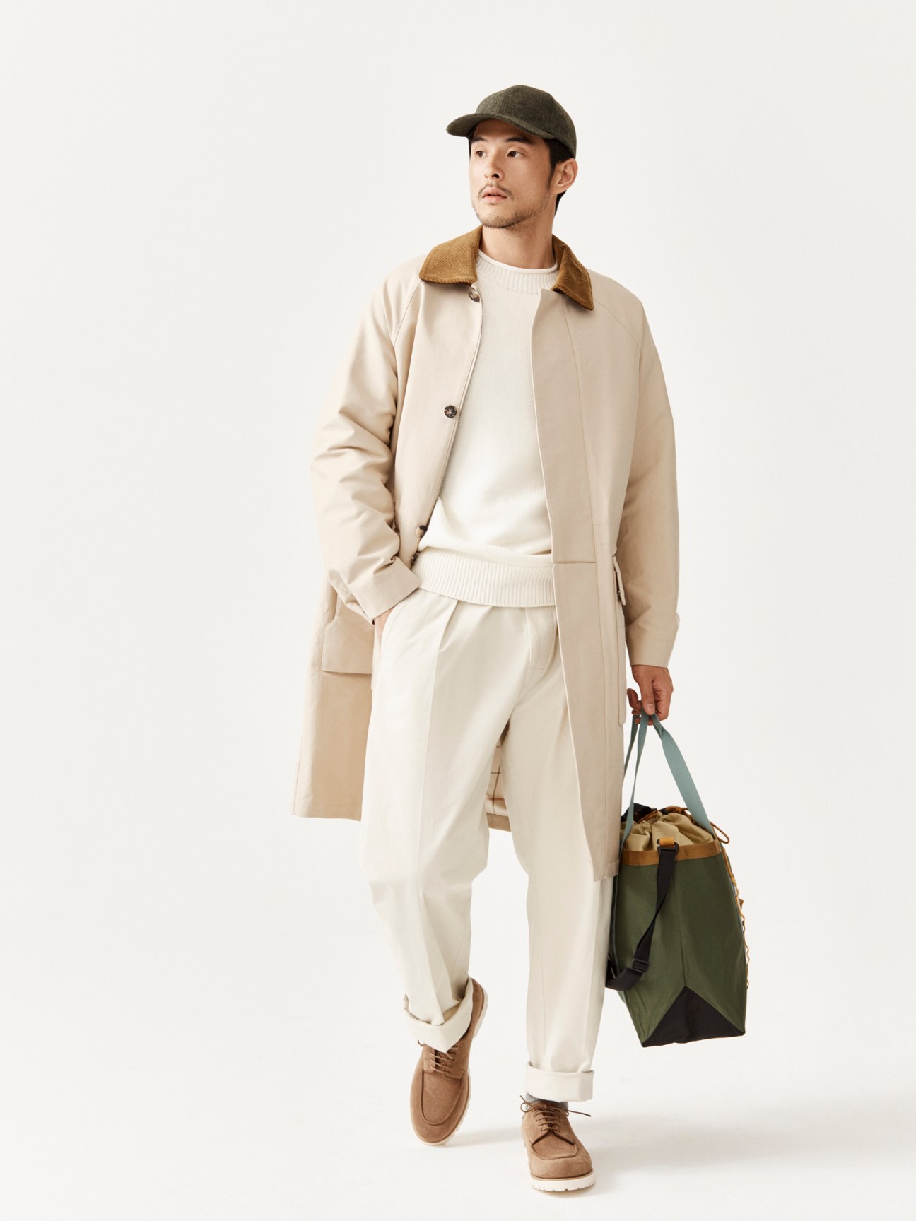 Man in beige trench coat and cream outfit holding green bag, wearing brown cap against white background