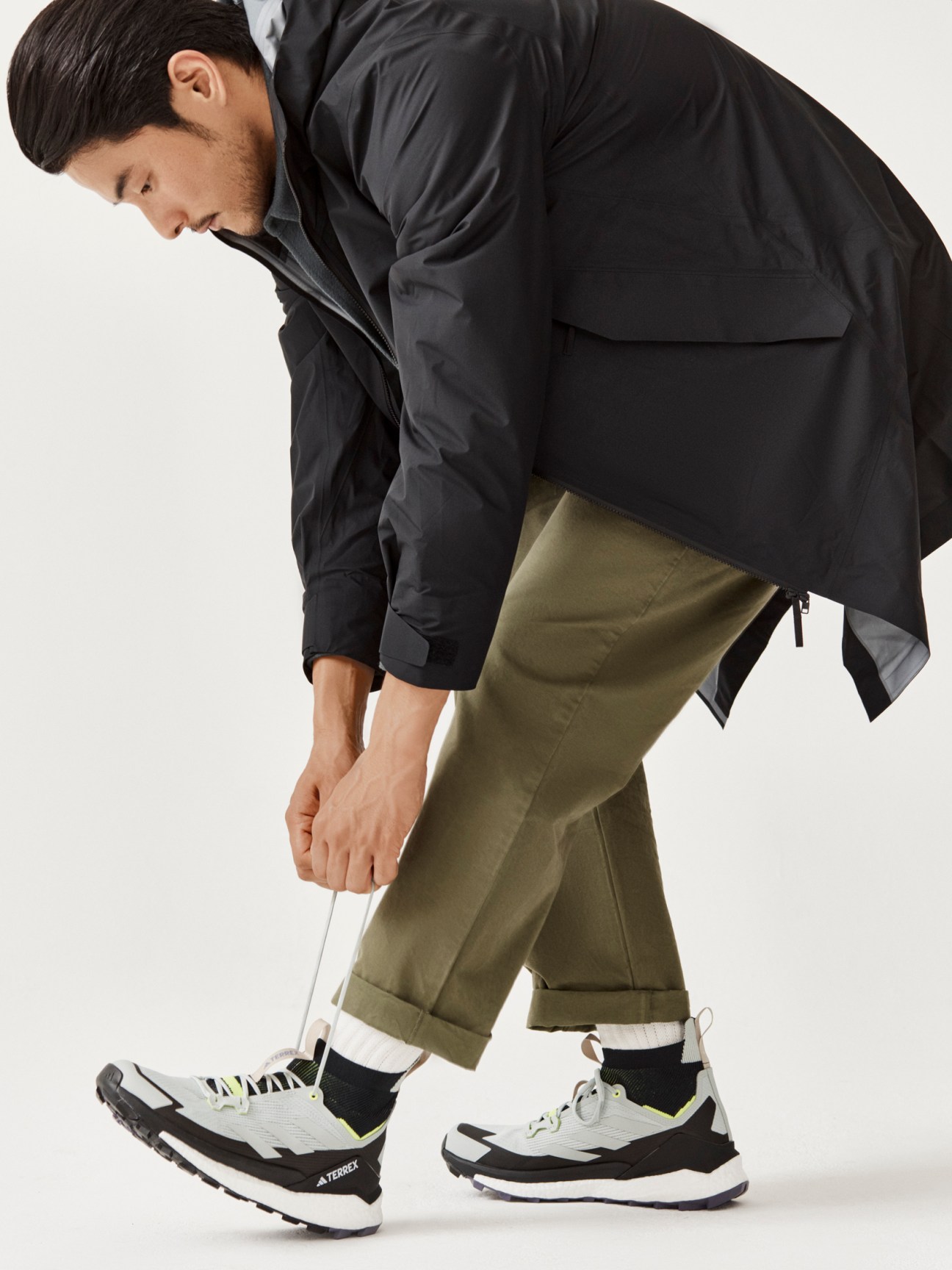 Man in black technical jacket and olive pants bending down to tie athletic sneakers against white background