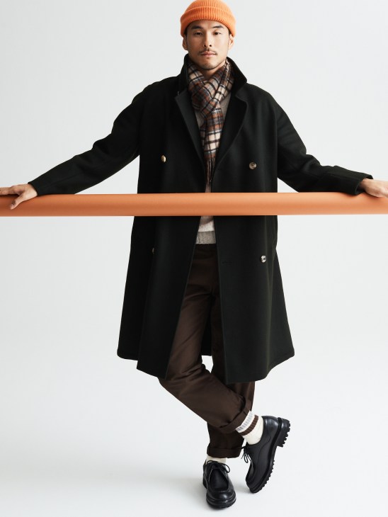 Model wearing navy coat, cream jumper, dark trousers, beanie and scarf in outdoor setting